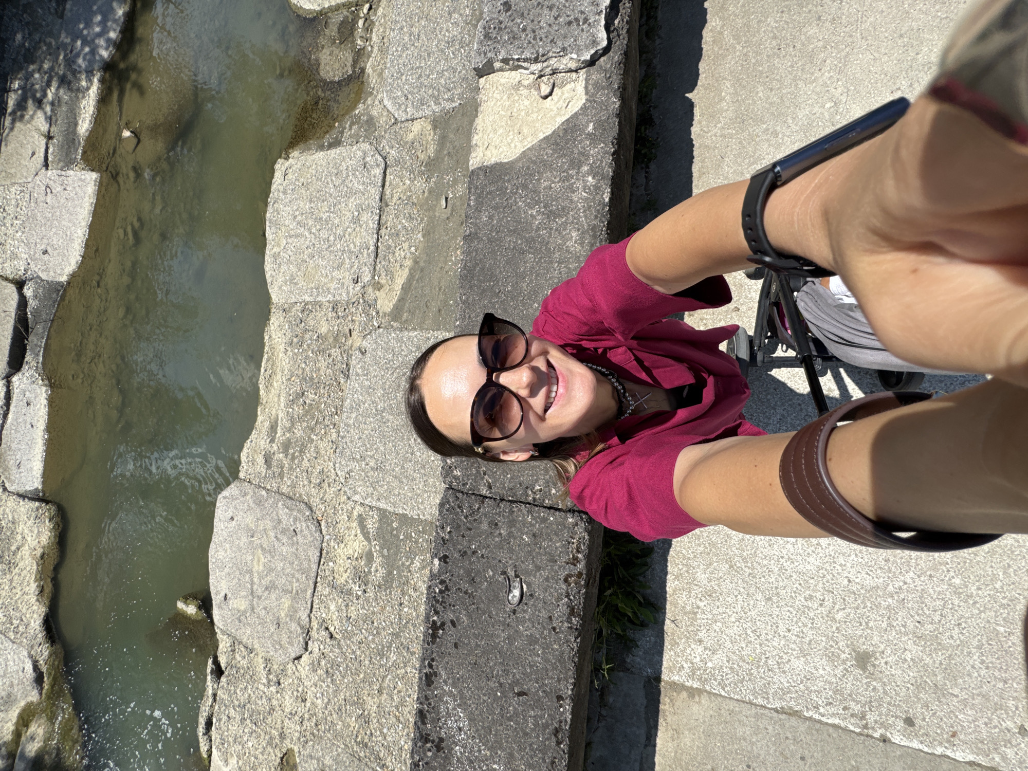 This is a selfie taken by a woman outdoors. She is smiling and wearing sunglasses and a magenta shirt. Her right arm is extended, holding the camera. A stroller is visible behind her. The setting appears to be a paved area next to a stone structure and a shallow body of water. The time of day is likely daytime, as indicated by the bright sunlight and shadows. The weather appears to be pleasant. No visible text is present in the image. The location context is Sochi, Russia.