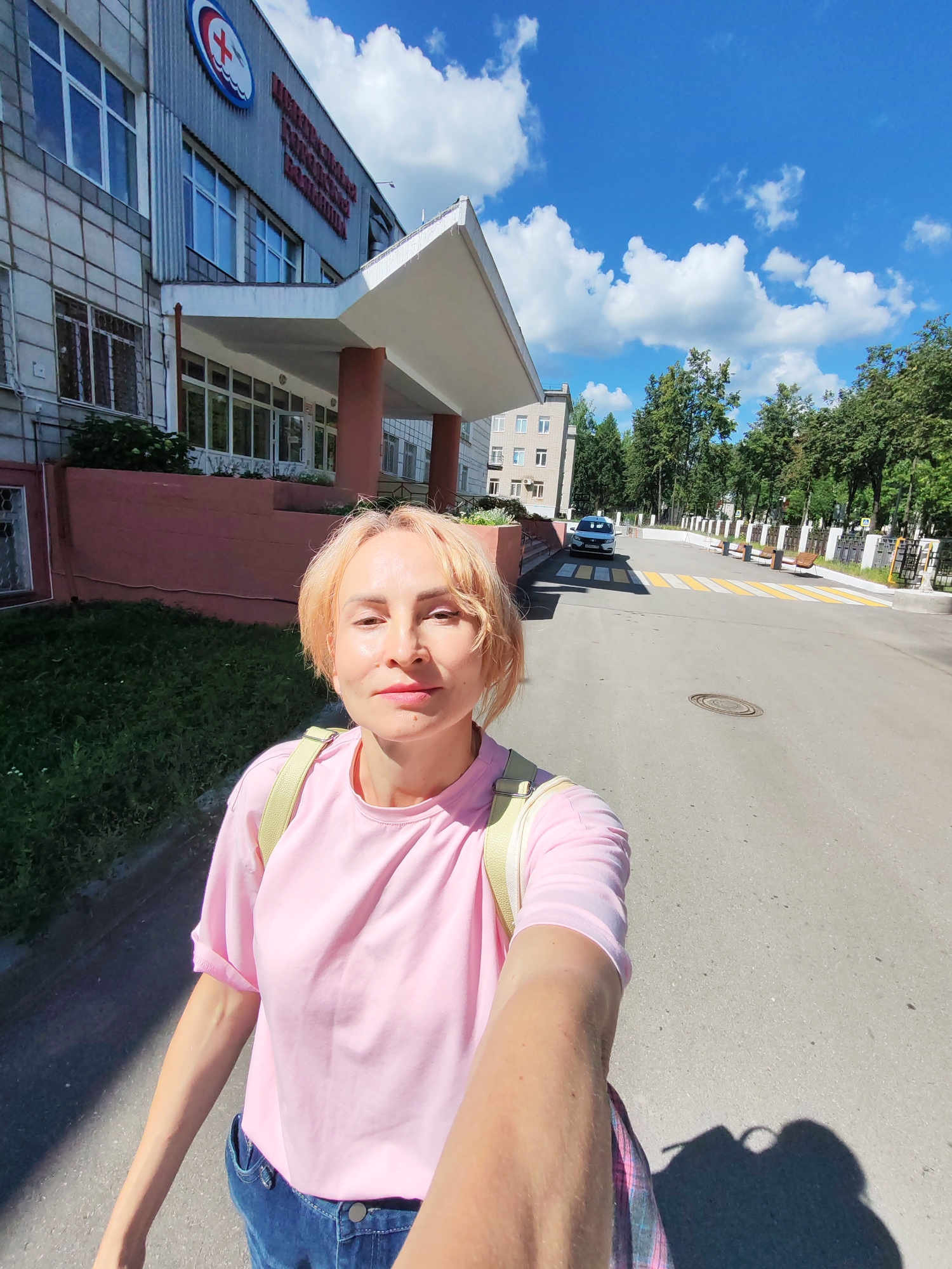A woman with short, blonde hair and fair skin is taking a selfie outdoors on a sunny day. She is wearing a pink t-shirt and blue jeans, with a light-colored backpack on her shoulders. In the background, there is a modern building with a red base and a white awning over the entrance, displaying a red cross logo. A street with a pedestrian crossing and benches runs alongside the building. Trees and other buildings are visible further down the street, and the sky is blue with white clouds. The building is identified as the Chaikovsky City Hospital.