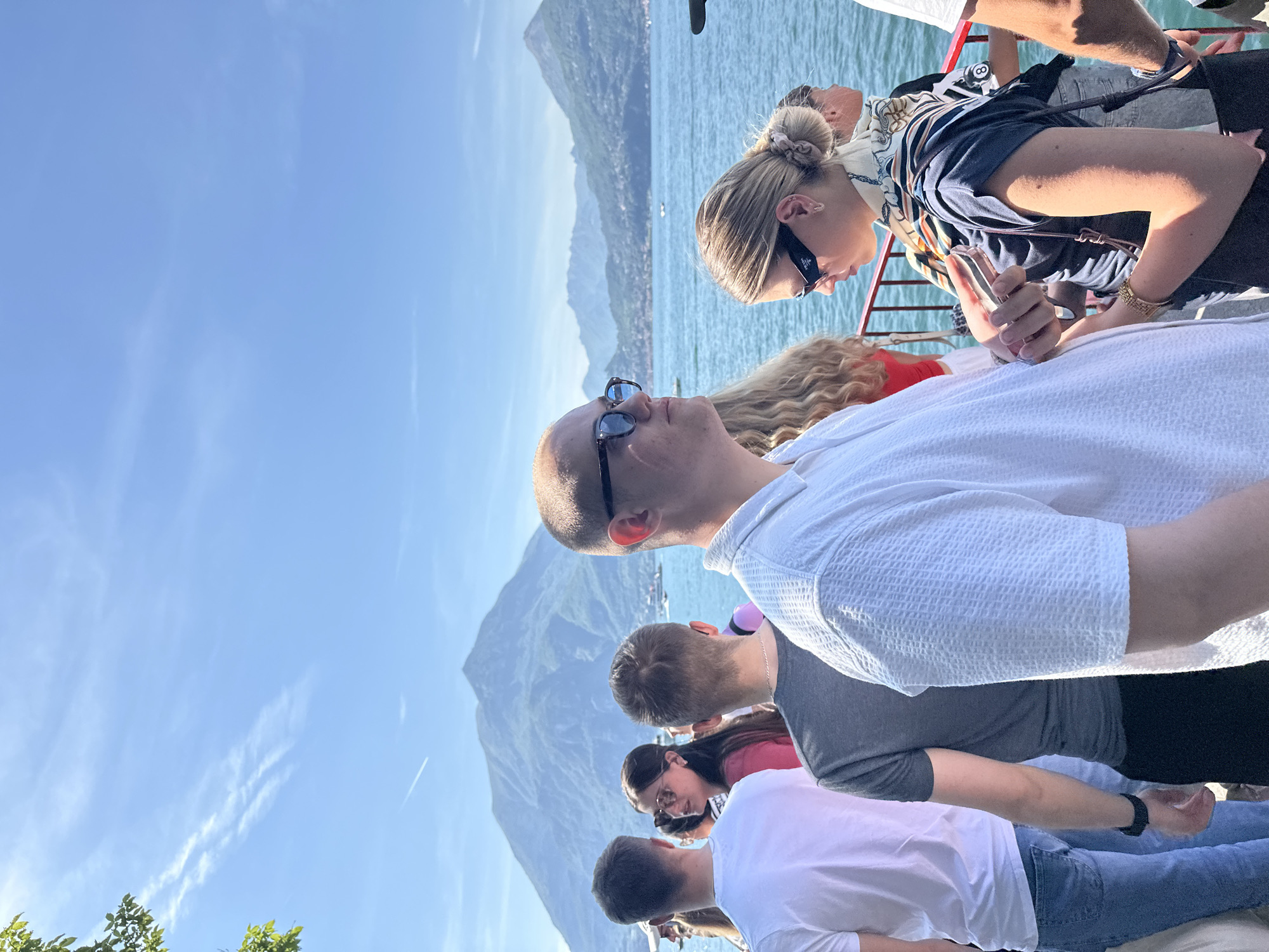 A daytime shot in Varenna, Italy, shows a group of people on a vessel. From left to right: A man in a white textured shirt and sunglasses looks up. Behind him, a man in a gray shirt stands next to a woman in a pink shirt and sunglasses. In the center, a woman in a navy top with a patterned scarf around her hair holds a phone. Behind her, a man in a white shirt and sunglasses leans over the railing. Mountains and water are visible in the background. The sky is blue with wispy clouds.