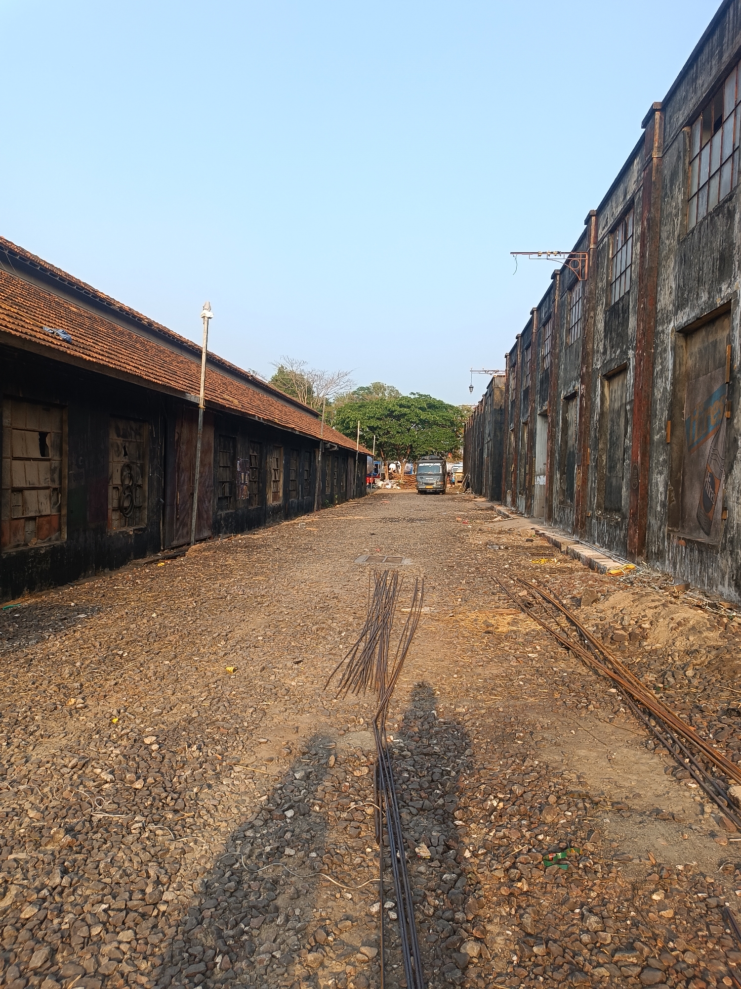 The image depicts a narrow, unpaved street or alley in what appears to be an industrial area or a decaying part of a city. The ground is covered in gravel and dirt, with rebar scattered haphazardly. On the left side is a long, low building with a tiled roof and dark, weathered walls, suggesting age and neglect. Windows are visible along the building's length, some broken or boarded up. On the right side stands a taller building with a concrete or stone facade. Its walls are stained and marked, and numerous windows are present, some boarded. In the distance, figures can be seen engaged in unknown activities near a vehicle. A canopy of green trees sits at the far end of the street, providing a point of natural contrast. The sky above is clear and light.