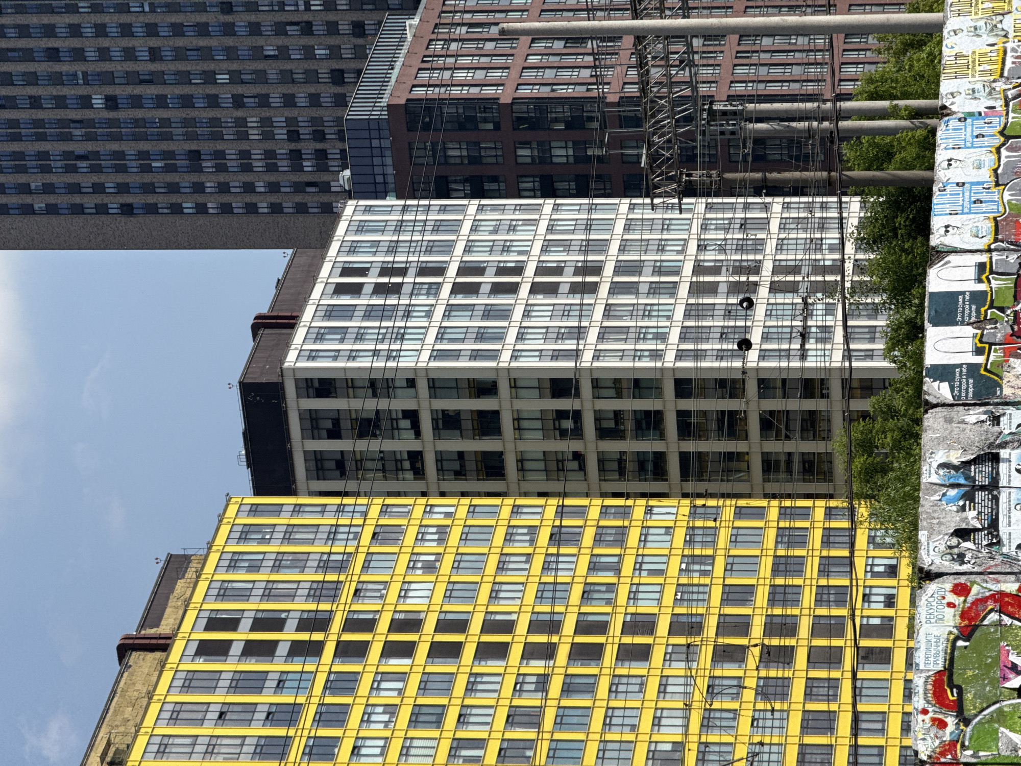 This is a medium-shot, vertical, exterior photograph taken on a sunny day in Khimki, Russia. The scene is dominated by several modern high-rise apartment buildings. On the left, a vibrant yellow building with a grid of numerous windows is prominently featured. To its right, a white building with a similar grid pattern of windows stands tall. Further to the right, a dark red brick building with many windows is visible, and behind it, a very tall, dark grey brick skyscraper rises into the clear blue sky. In the foreground, a graffiti-covered concrete wall is plastered with various posters and paper scraps. Power lines and poles are visible between the buildings and in front of them. Lush green trees are interspersed among the buildings and behind the foreground wall.