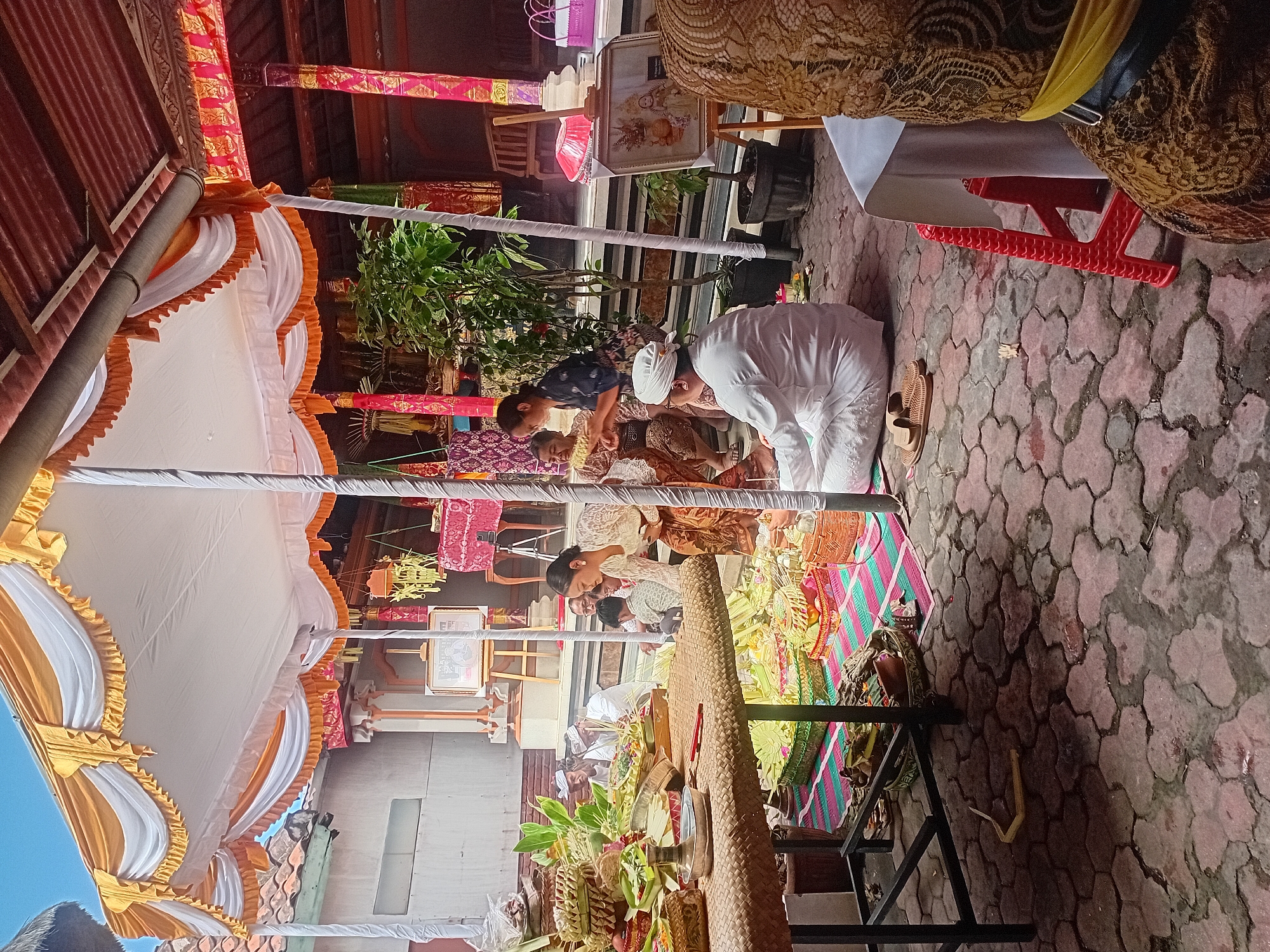 In Kuta, Indonesia, a Balinese ceremony is underway beneath a large, orange and white tent. The ground is paved with interlocking hexagonal bricks. Several individuals are gathered, some seated on the ground, others standing. In the foreground, a woven table is laden with offerings in woven baskets, consisting of fruits, vegetables, and leafy greens. A man dressed in a white traditional outfit, including a head covering, is seated on the ground. Several other people in traditional Balinese attire are also present. Behind them, a red stool and a table draped with a white cloth are visible. The environment appears to be an outdoor courtyard, possibly part of a temple or traditional Balinese home, with structures and decorative elements suggestive of the local culture.