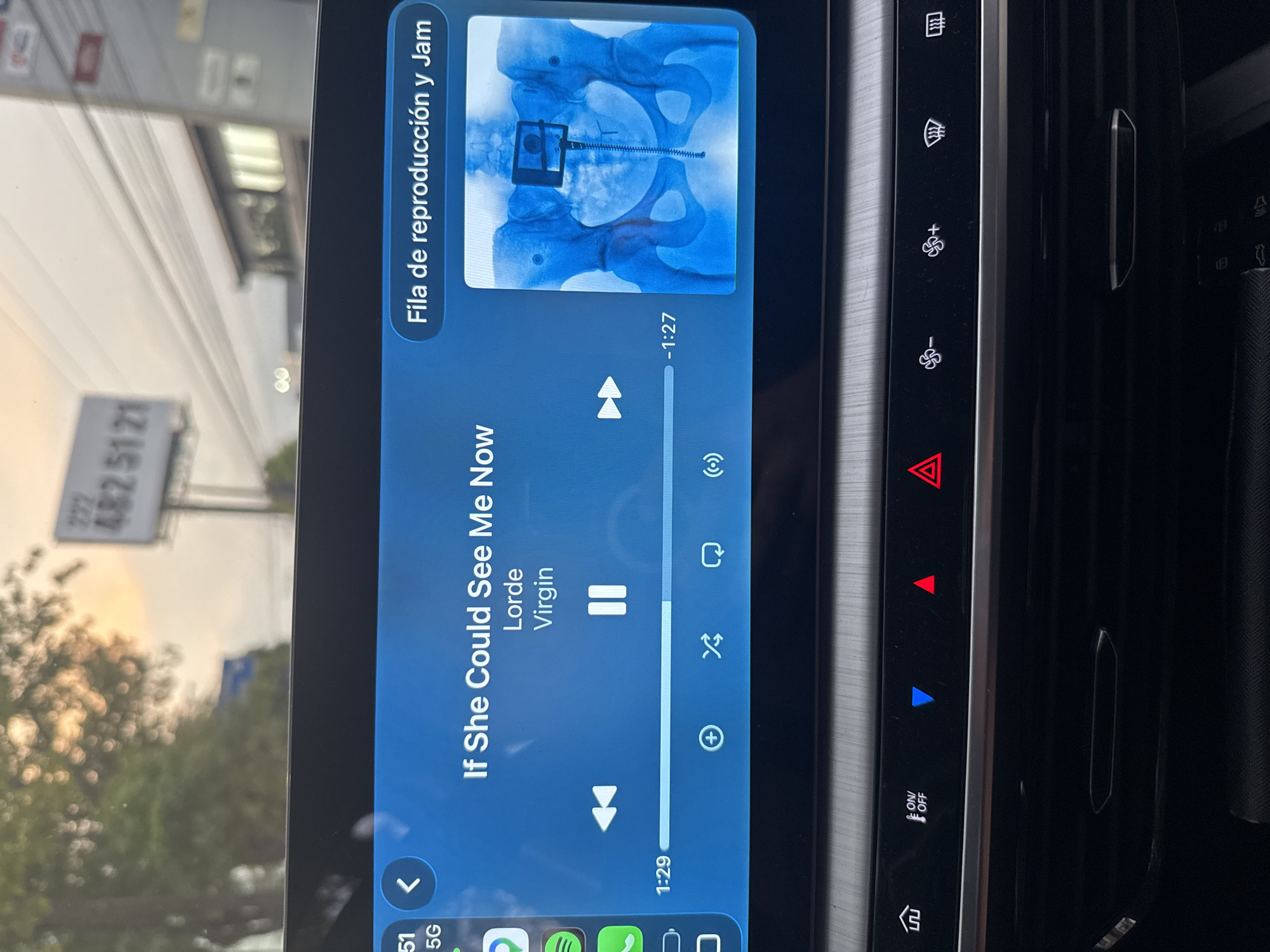 The image shows a car's infotainment screen displaying music playback information. The song "If She Could See Me Now" by Lorde from the album "Virgin" is playing. The cover art is an X-ray of a pelvis with surgical hardware. The screen indicates the song is 1 minute and 29 seconds long and has 1 minute and 27 seconds remaining. Adjacent to the screen are car control buttons including defrosters, climate control, hazard lights, and an on/off button. The background shows an exterior view of a street with buildings and trees; a sign with the number 482 5121 is visible, suggesting a location in or near Puebla, Mexico. The phone's signal is 5G.