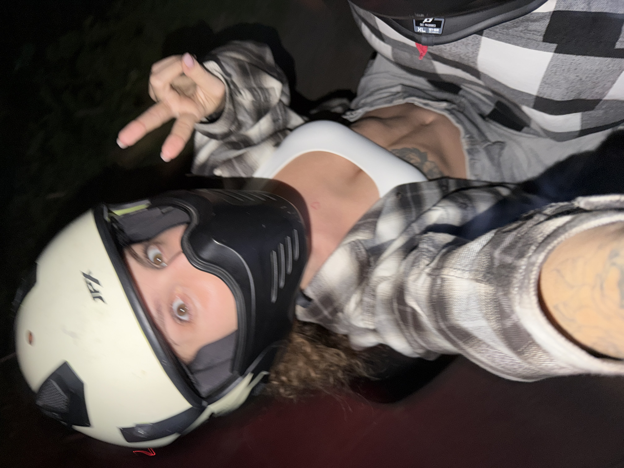 The image captures a sideways portrait of a person seated on what appears to be a motorcycle or scooter at night, likely in North Kuta, Indonesia. The subject, an individual with curly brown hair, wears a cream-colored motorcycle helmet, a white sports bra, and an unbuttoned black and white plaid long-sleeve shirt. Their wide-open brown eyes gaze directly at the camera, and one hand is raised to form a peace sign, revealing light pink nail polish. Visible tattoos include a small red heart on the lower abdomen and an intricate design on the arm. The helmet features "JPL" text on its side, and the plaid shirt's inner collar tag shows "DC PASSED" and "XL". The background is dark and blurred, indicating a low-light, outdoor setting. The pose and expression suggest a playful or excited mood.