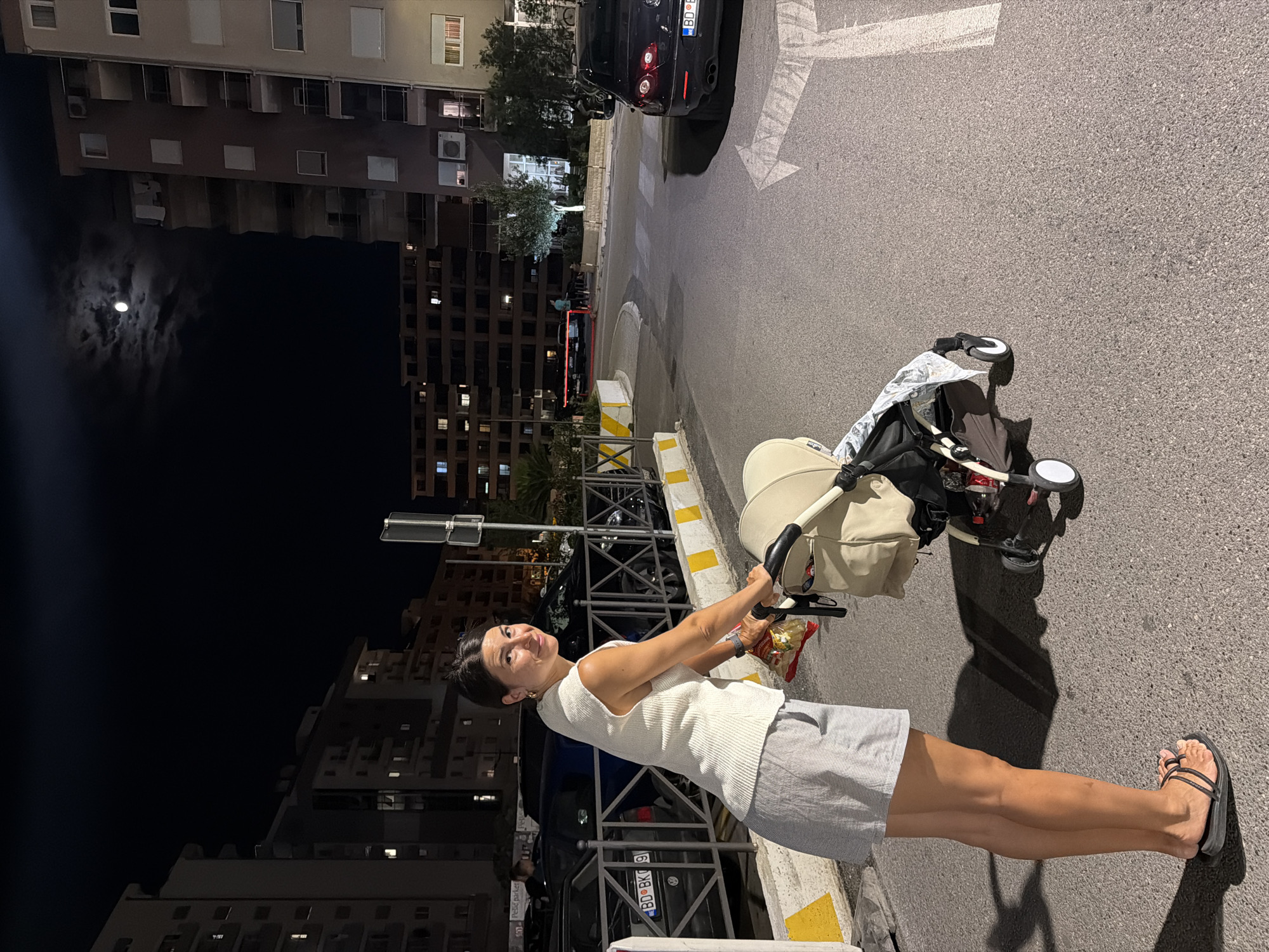 A full-body shot features a woman dressed in a white sleeveless top and a grey and white striped skirt, pushing a baby stroller. She is facing the camera and smiling. The stroller is cream-colored with a grey canopy and has some items inside. The woman is wearing black sandals. She is holding a package of what appears to be pasta in her right hand. The scene is outdoors at night, with the moon visible in the dark sky. Tall buildings with illuminated windows are in the background, suggesting an urban environment. The ground is asphalt with painted white lines, indicating a street or parking area. A black car is parked in the background. The overall mood is casual and pleasant. The license plate of the car is visible and reads "BD BK91".