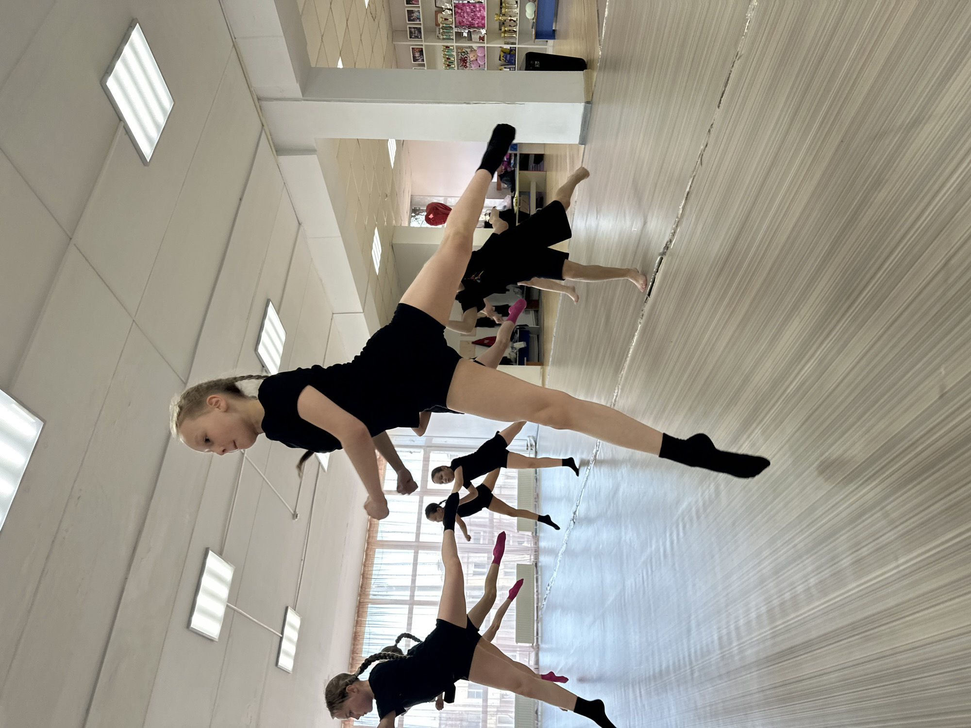 A wide-angle shot depicts a dance class of young girls in Votkinsk, Russia. The interior features a light, airy gymnasium with white walls, fluorescent lighting panels, and a light wood-grain floor. There are approximately six girls, all wearing black shorts and tops. They are performing a synchronized exercise, with one leg extended upwards, the other angled towards the floor, and arms held in a similar position. Some wear black socks, while others wear pink. In the background, there is a shelf with trophies and decorative items, suggesting a history of athletic success. The scene suggests an active dance or gymnastics practice.