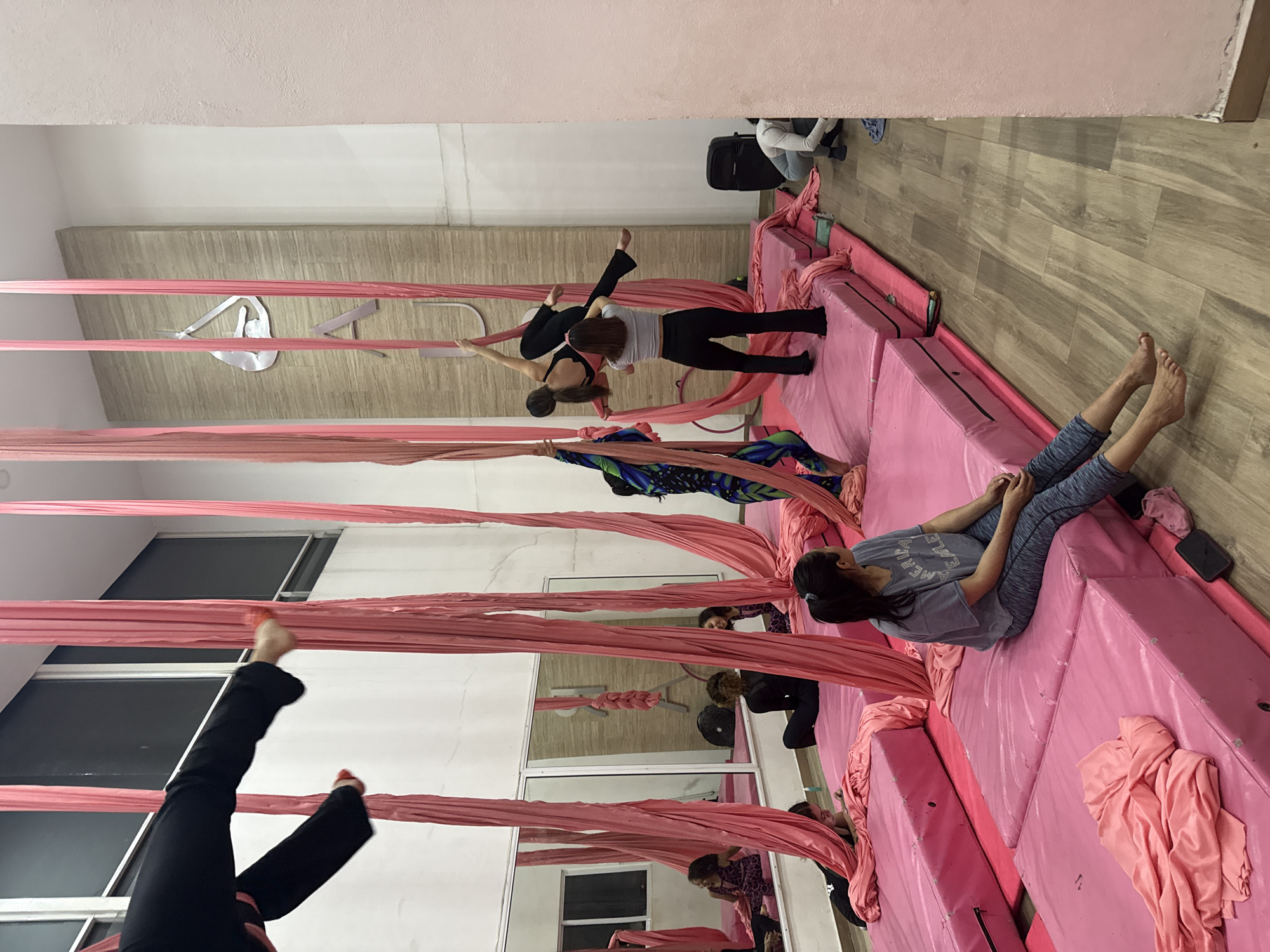 An indoor aerial silks studio in San Andrés Cholula, Mexico, features multiple participants engaged in or resting from practice. The room has high white walls, a light-colored wooden floor, and large pink safety mats covering the floor beneath the hanging silks.
Several sets of vibrant pink aerial silks hang from the ceiling. In the foreground, one person, dressed in black, is upside down in an inverted pose on the silks. Towards the mid-ground, two individuals are interacting near another set of silks; one in a white top and black pants appears to be assisting or spotting another person in a complex aerial pose on the fabric. To the right, a person with dark hair, wearing a grey t-shirt and patterned grey leggings, is seated on the pink safety mats, appearing to rest. The text "AMERICA EMBLEM" is partially visible on their shirt. In the background, another person is partially visible near a black bag.
Mirrors along one wall reflect parts of the studio and the participants, adding depth to the scene. On a far wall, a stylized metal sign features a figure in an aerial pose, with letters, including a clear 'A' and part of a 'D', visible next to it. The studio is well-lit, suggesting either natural light or bright artificial lighting. The overall atmosphere appears focused and active, typical of a training session.