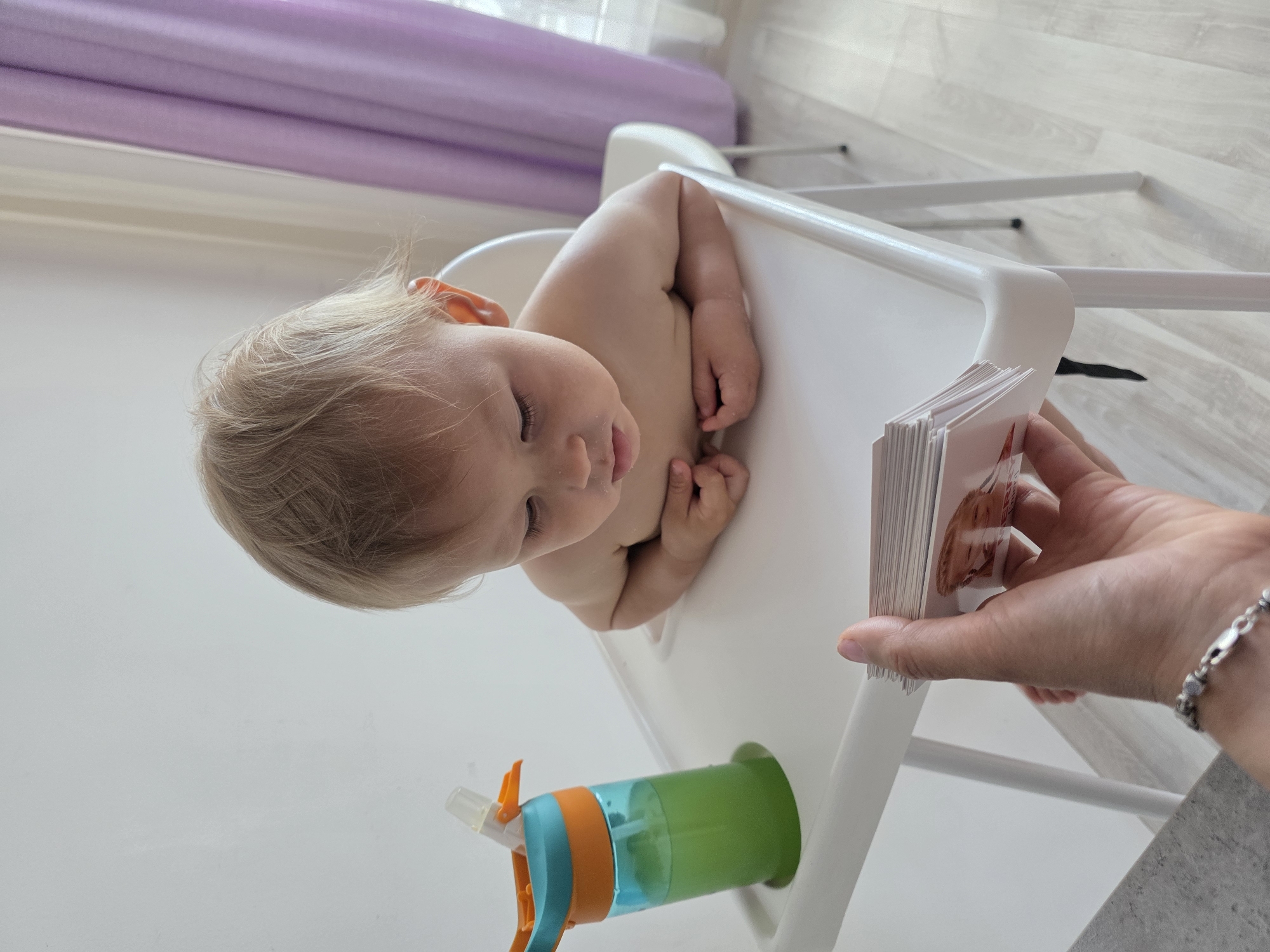 A young child with blond hair sits in a white high chair, looking down at a stack of cards held by a person's hand. The child is shirtless and has some powder around their mouth. To the left of the child is a blue and orange sippy cup filled with a green liquid. The background shows a white wall and purple curtains. The scene appears to be indoors.