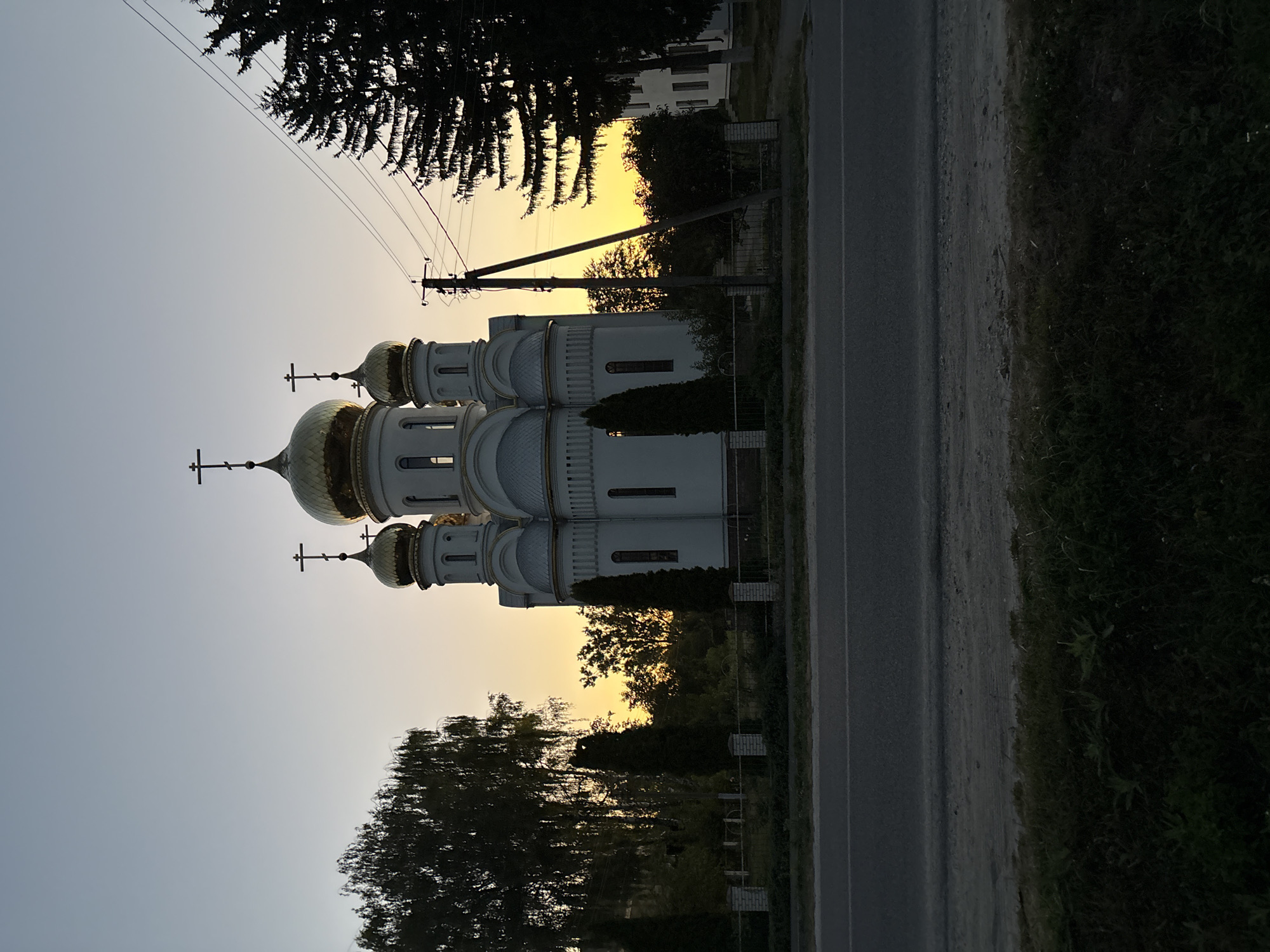 The image depicts a white church with golden domes against a dusky sky. The church is surrounded by trees and a fence. A road is visible in the foreground, with grass and dirt on the side. Utility poles and power lines are also present in the scene. The overall atmosphere is serene, possibly captured during sunset or sunrise. The location is the city of Lanivtsi, Ukraine.