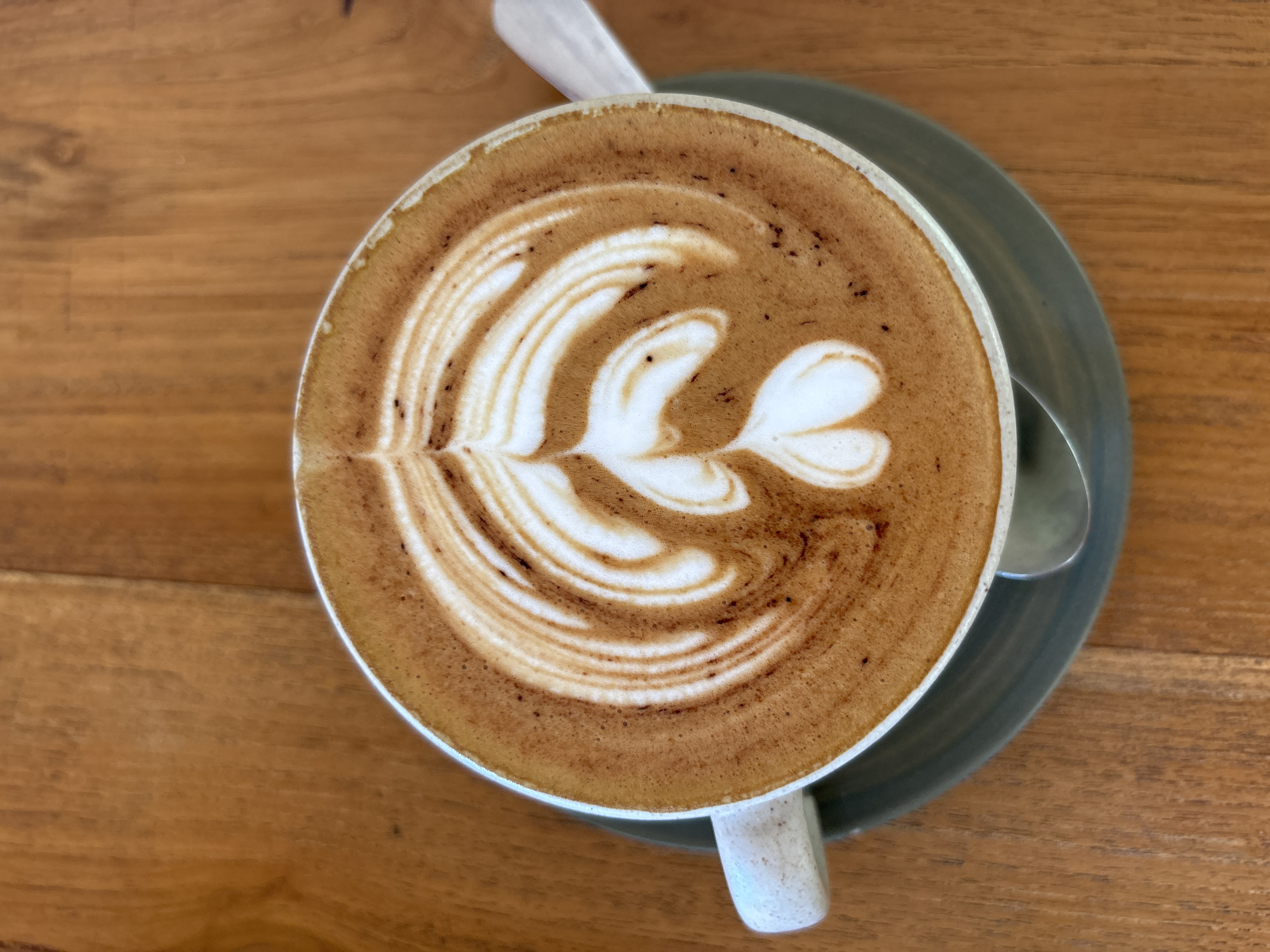 A top-down, close-up shot reveals a cup of latte art on a wooden table. The coffee cup, containing a creamy brown beverage with a white foam design resembling a leaf and hearts, rests on a saucer. A small spoon sits beside the cup on the saucer. The table exhibits a light-brown wood grain. The image was likely taken indoors in Kuta Selatan, Indonesia, given the prompt.