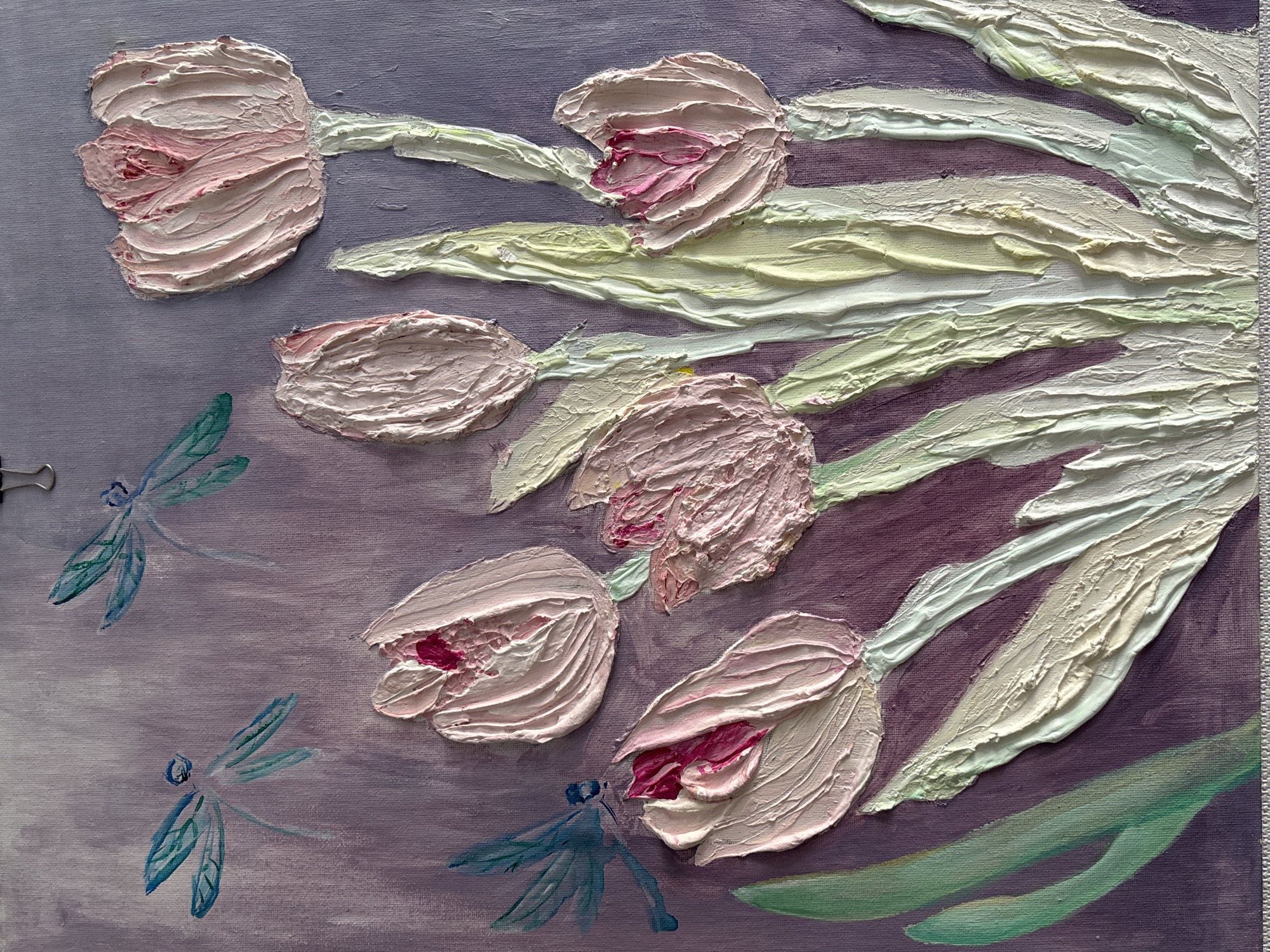 The image is a close-up, high-angle shot of an impressionistic painting of tulips and dragonflies. The painting uses thick impasto strokes, giving the flowers and leaves a textured, almost sculptural quality. The tulips are predominantly light pink with touches of darker pink and red inside the petals. The stems and leaves are rendered in shades of light green and cream. Three teal-colored dragonflies are depicted in flight, with translucent wings. The background is a soft, textured lavender. The painting is on canvas, with the texture of the canvas visible. The top edge of the canvas shows a metal clip holding it in place. There is no information available that can locate this image in the city of Pavlodar, Kazakhstan.
