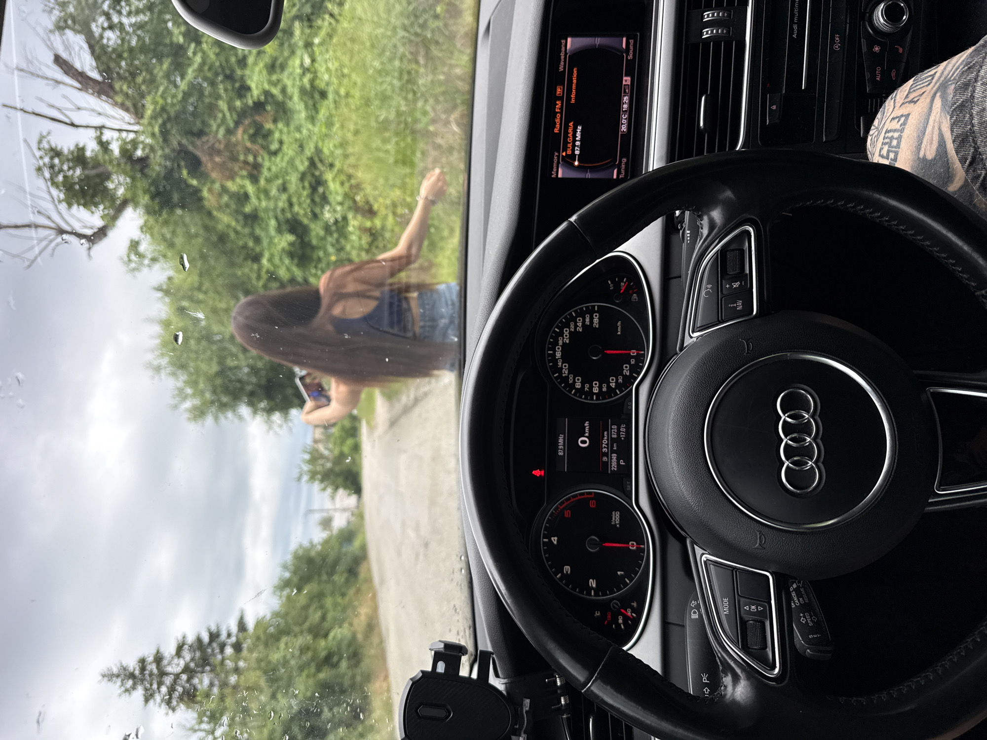 An eye-level, medium shot captures the interior of a car, looking through the windshield at a woman standing on a dirt road in a rural area. The car's dashboard, steering wheel, and a phone mount are in the foreground. The woman, with long brown hair, is holding a smartphone and appears to be taking a picture or recording a video. She is wearing a blue bra-top and denim shorts. The background consists of lush green trees and a cloudy sky. The car's infotainment system is visible, displaying the radio station "BULGARIA" and the time "18:25". The road is unpaved and appears to be in a state of disrepair. The overall scene suggests a spontaneous stop during a drive.