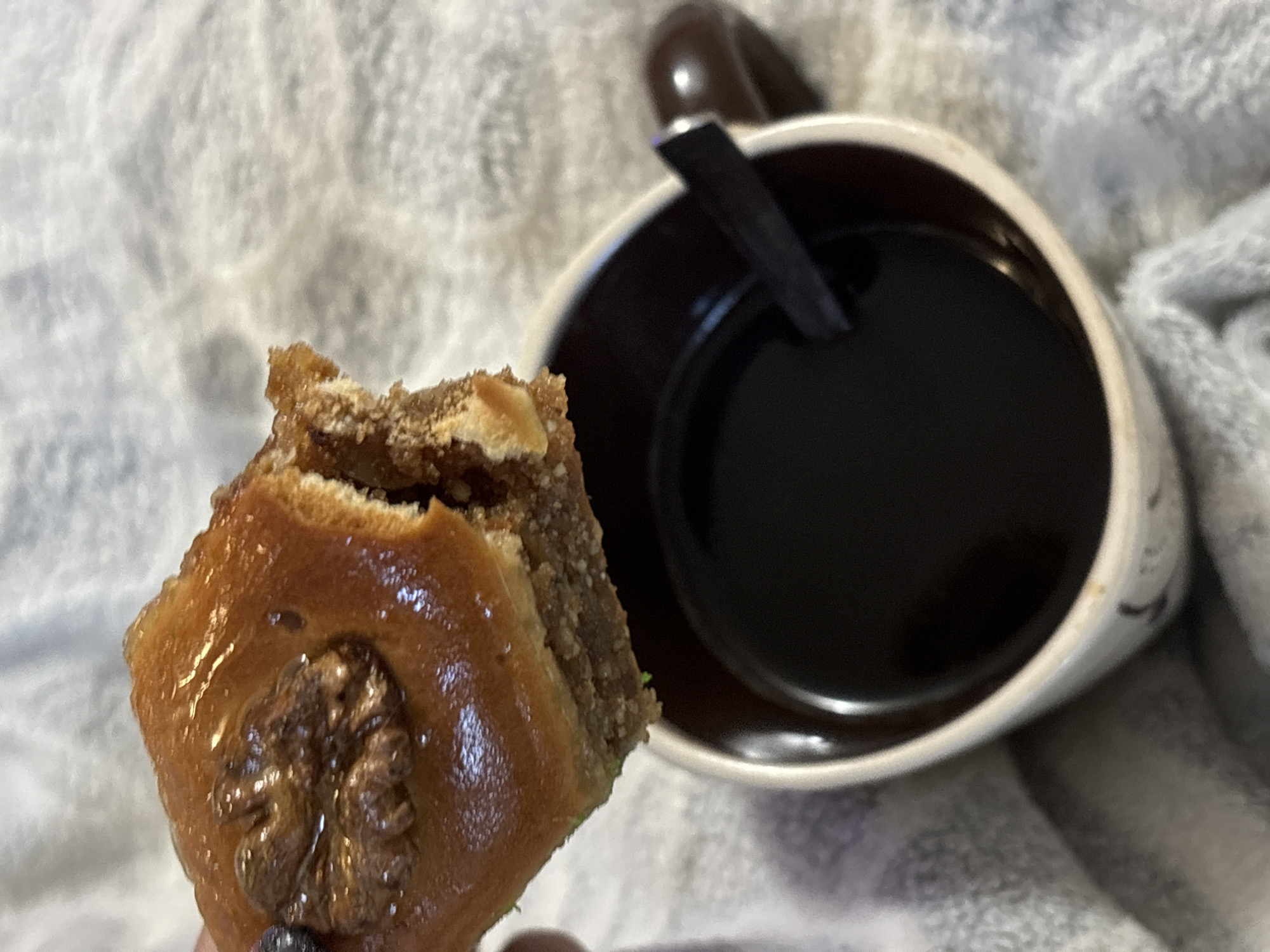 A close-up shot shows a piece of baklava held above a mug of dark coffee, resting on a grey, textured blanket. The baklava is golden brown and appears to be drizzled with syrup, with a walnut embedded in its top layer. A bite has been taken out of the baklava. The mug is white with a dark brown rim and handle. A dark, flat stirring utensil is partially submerged in the coffee. The image is shot from a slightly elevated angle, focusing on the food and drink. There are no identifiable landmarks or elements that would suggest a specific location within Chaikovsky, Russia.