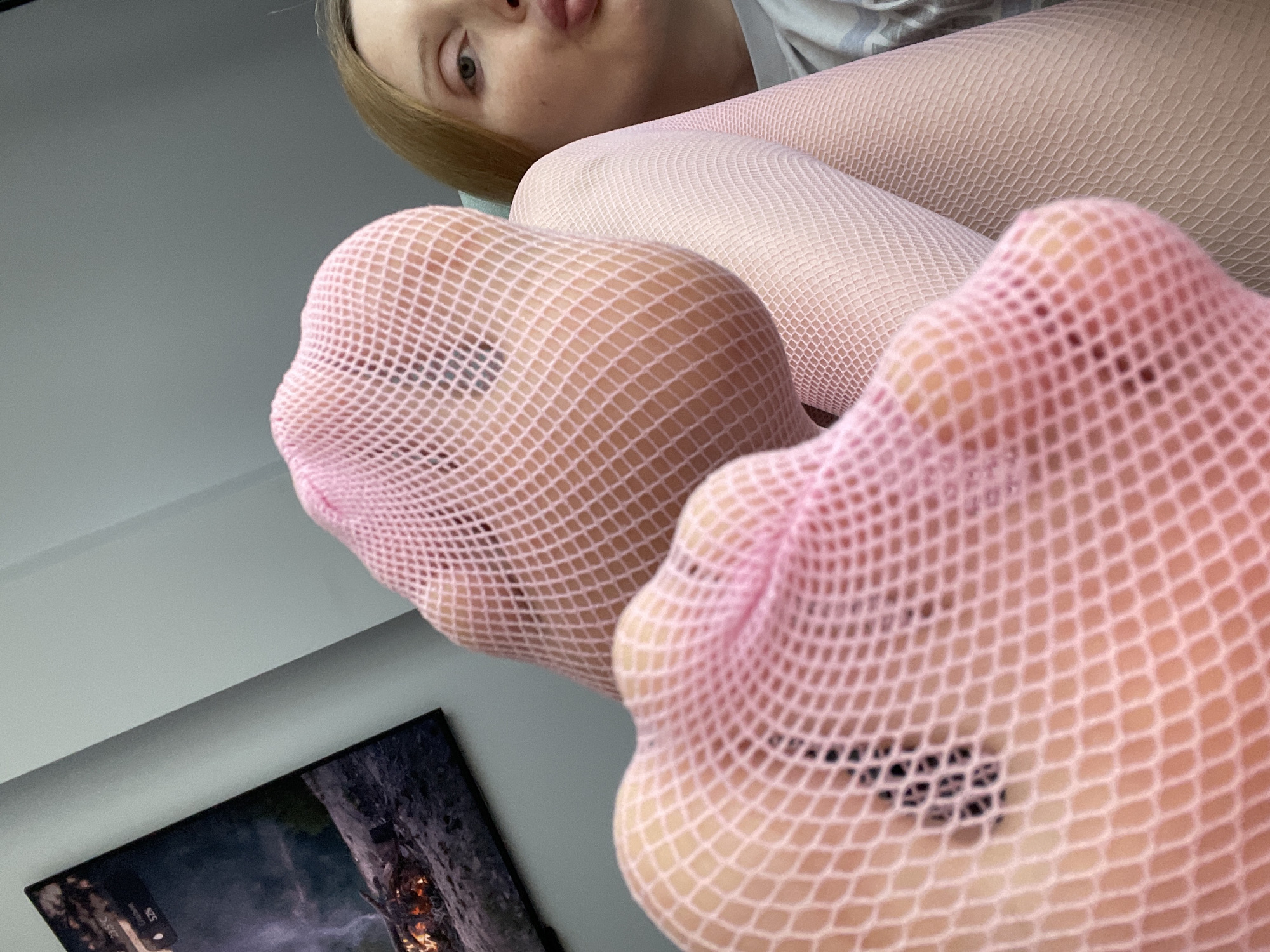 The image is a close-up, low-angle shot of a person's feet, wearing pink fishnet tights. The feet are positioned in the foreground, with the toes of both feet clearly visible through the mesh. The person's face is also visible in the upper portion of the image, looking down at the camera with their tongue sticking out slightly. They have blonde hair.
The background appears to be an indoor setting with a gray wall. To the lower left, a portion of a dark-colored screen displaying an image with fiery elements is visible, suggesting the presence of a television or computer monitor. The lighting suggests it is daytime, possibly indoors with artificial lighting.
The overall tone of the image is playful and intimate, focusing on the textures of the fishnet and the person's playful expression. There are no discernible signs of weather or specific time of day beyond general indoor lighting. The location context provided is Yablonovskiy, Russia. No text is visible in the image.