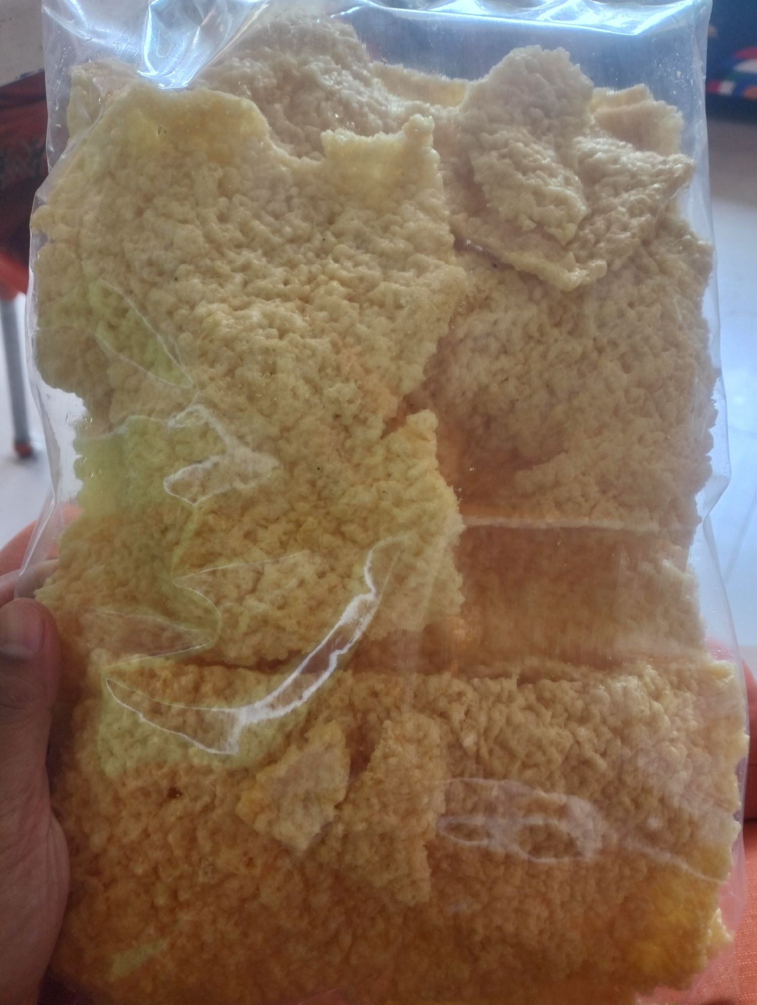 A person's left hand is visible on the left side of the image, holding a clear plastic bag filled with light yellow, porous, and irregularly shaped food items. The food appears to be a snack, possibly a type of cracker or crispbread, stacked inside the bag. The background is blurred, but suggests an indoor setting with hints of furniture and possibly other objects. The location is identified as Air Jamban, Indonesia, though no specific landmarks or contextual clues in the image directly support this location.