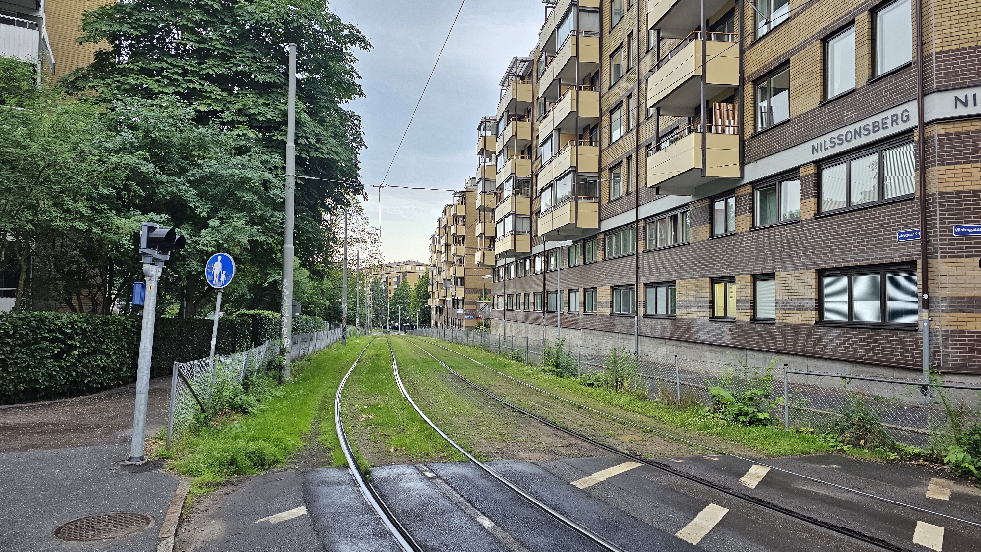 This image shows a street scene in Gothenburg, Sweden. The foreground features a set of tram tracks curving to the left, bordered by a paved path and a low concrete wall. To the left of the tracks, a traffic light signal with three lights (two black and one red) stands tall, accompanied by a blue circular sign depicting a person walking with a child. A tall, thin lamppost is situated further down the path.
The background is dominated by a long, multi-story apartment building with a distinctive brick facade of alternating yellow and brown bands. The building has numerous windows and balconies with light yellow railings. The name "NILSSONSBERG" is prominently displayed on the building's facade, along with street signs indicating "Världgatan 94" and "Världgatan". A fence runs along the edge of the tram tracks, separating them from the building and a dense line of green bushes and trees.
On the left side of the image, a large tree with lush green foliage overhangs the scene, providing shade. The tram tracks themselves are lined with green grass, suggesting a well-maintained urban environment. In the distance, more apartment buildings and trees are visible, hinting at a residential area. The sky is overcast, suggesting a cloudy day. There are no people or active events depicted; the scene appears to be a static view of an urban streetscape.