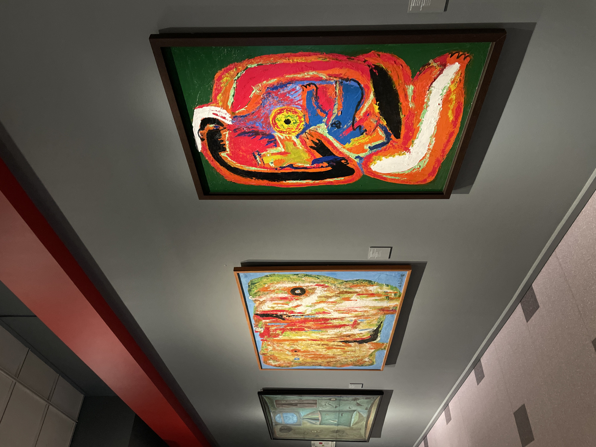 This image depicts an interior view of an art gallery, specifically identified as the New Tretyakov Gallery in Moscow, Russia. No individuals are present in the scene.
Three framed abstract paintings are displayed on a dark grey wall. The most prominent painting, positioned on the right, is a large, vertically oriented work. It features a vibrant green background and a central, abstract figure rendered with thick, impasto brushstrokes in bright red, orange, black, white, blue, and yellow, highlighted by a distinct yellow eye-like circle at its core. This artwork is enclosed within a dark brown frame. To the left of this, a horizontally oriented abstract painting, enclosed in a light wooden frame, presents a textured composition primarily in shades of orange, yellow, red, green, and white against a light blue background, with a notable dark vertical line and two smaller dark circular forms. Further to the left, a smaller, dark-framed, vertically oriented painting exhibits abstract or surrealist forms predominantly in muted blue-green tones. Small white informational labels are affixed to the wall next to or below the artworks.
Above the displayed art, a wide, rectangular red beam extends horizontally along the upper portion of the grey wall. The floor is a light-colored, possibly pinkish-grey, surface with visible square patterns. A white tiled ceiling is partially visible in the upper-left quadrant of the image.