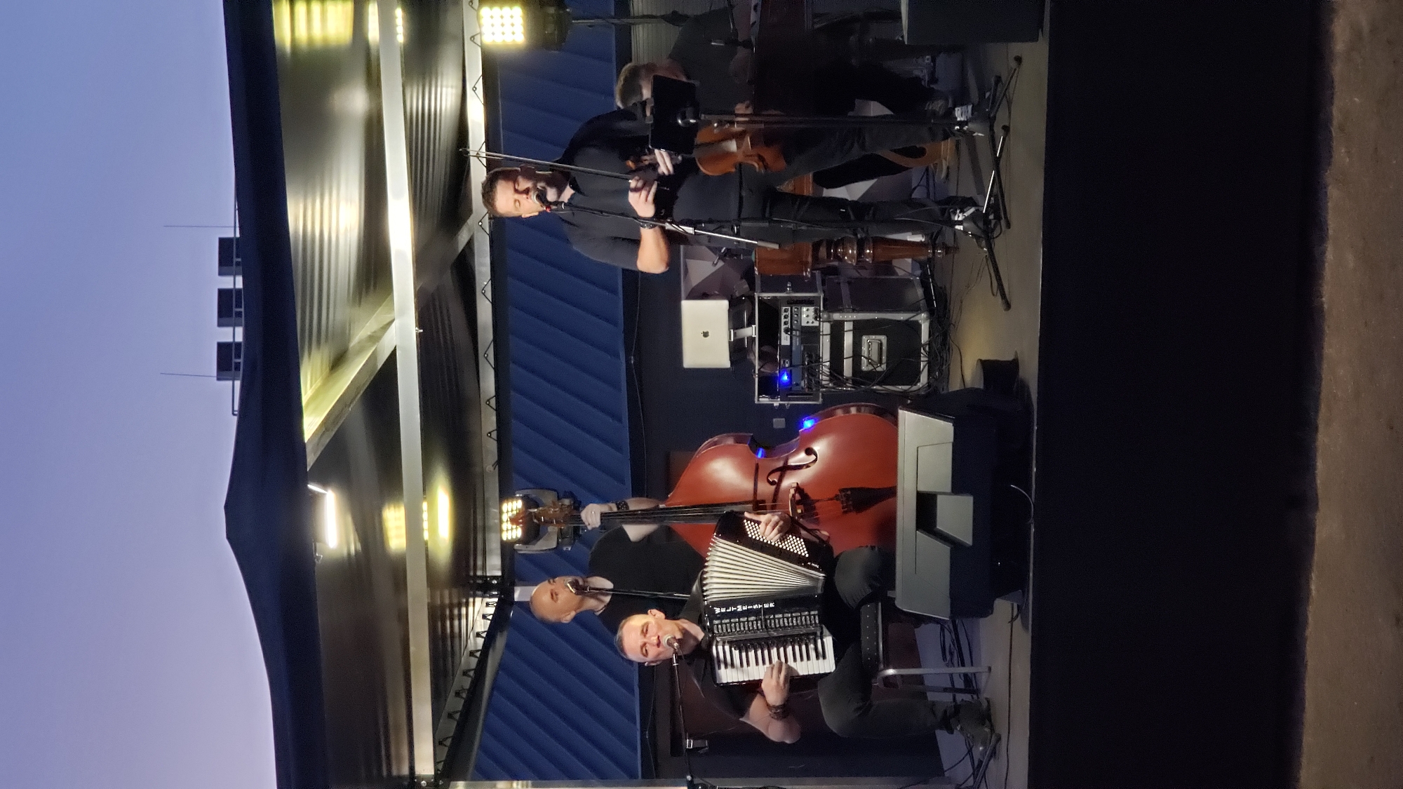 The image captures an outdoor evening performance on a stage under a canopy. Four men are actively engaged in playing music. On the left, a man sits on a stool playing a black accordion and singing into a microphone. To his right, a man stands behind a large, reddish-brown double bass, also singing into a microphone. In the center, a man stands playing a violin and singing. To the far right, another man is partially visible, seated and appearing to play a keyboard instrument. The stage is equipped with various sound equipment, including speakers, mixers, and monitors, along with lighting fixtures illuminating the performers. The background shows a dark blue structure, possibly part of a building, against a twilight sky. The overall setting suggests a concert or musical event. While the location is stated to be Prešov, Slovakia, there are no specific visual cues within the image that confirm this exact location.