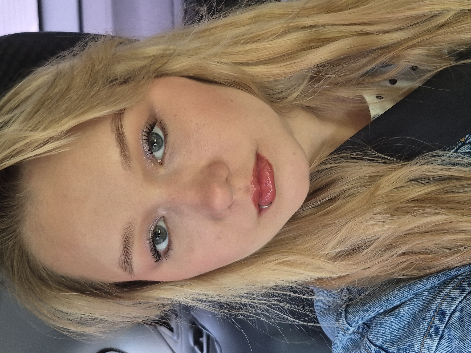 A young woman with long blonde hair and blue eyes is photographed from the chest up. She is wearing a denim jacket over a light-colored shirt with black polka dots, and a black seatbelt across her chest. She has makeup on, including eyeshadow and lip gloss. She has a lip ring in her lower lip. The background appears to be the interior of a vehicle, possibly a car, with dark seats visible on the left. The lighting suggests an indoor or shaded outdoor setting. There is no discernible information in the image that would indicate a location in Chaikovsky, Russia.