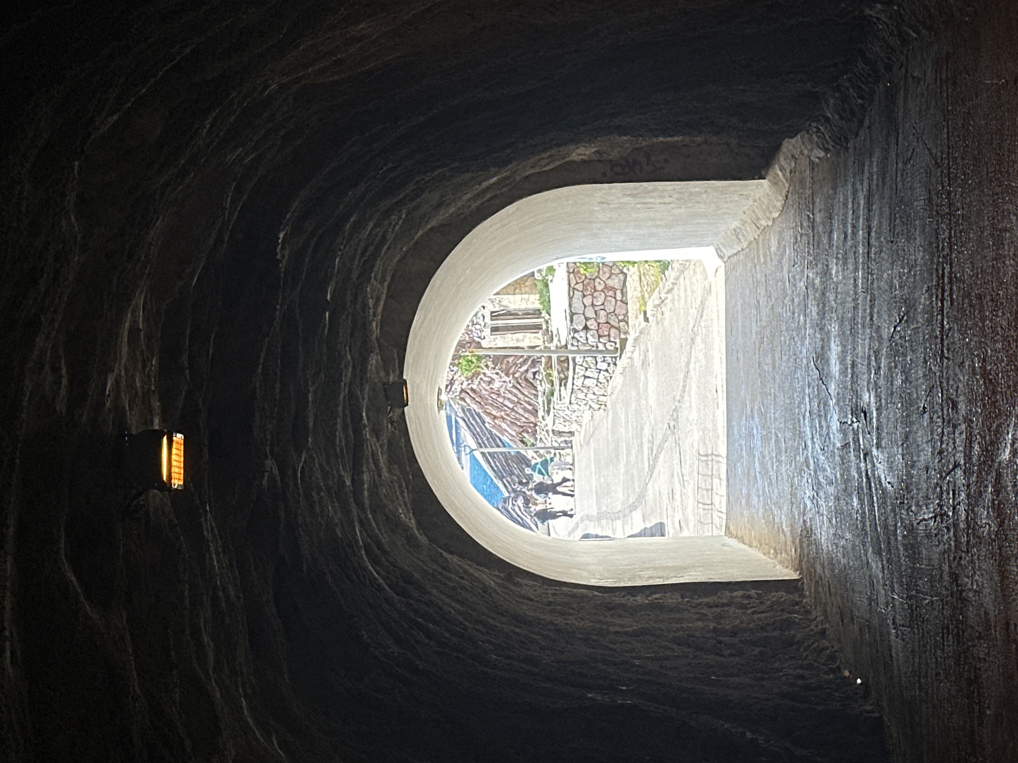 The image depicts a perspective from a dark, textured interior looking out through a bright, semi-circular archway. The immediate foreground is dominated by the rough, undulating surfaces of a cave or tunnel wall, cast in deep shadow. On the left wall of this dark space, a small, rectangular light fixture emits a warm, orange-yellow glow.
The archway itself is made of a lighter, smoother material, possibly concrete, and provides a stark contrast to the dark interior. Through this bright opening, an outdoor scene is visible. Several indistinct figures, appearing to be people, are seen in the distance, standing or walking on a stone-paved path or steps. The exterior features various light-colored architectural elements, including walls made of stacked stones, concrete surfaces, and hints of green vegetation. A splash of blue, possibly a railing or water, is also discernible.
The dramatic light contrast between the shadowy interior and the brightly lit exterior suggests it is daytime outside. The scene evokes a sense of looking out from a sheltered or hidden space into an open, sunlit environment, consistent with a Mediterranean coastal setting like Kamenovo, Montenegro. No visible text is present.