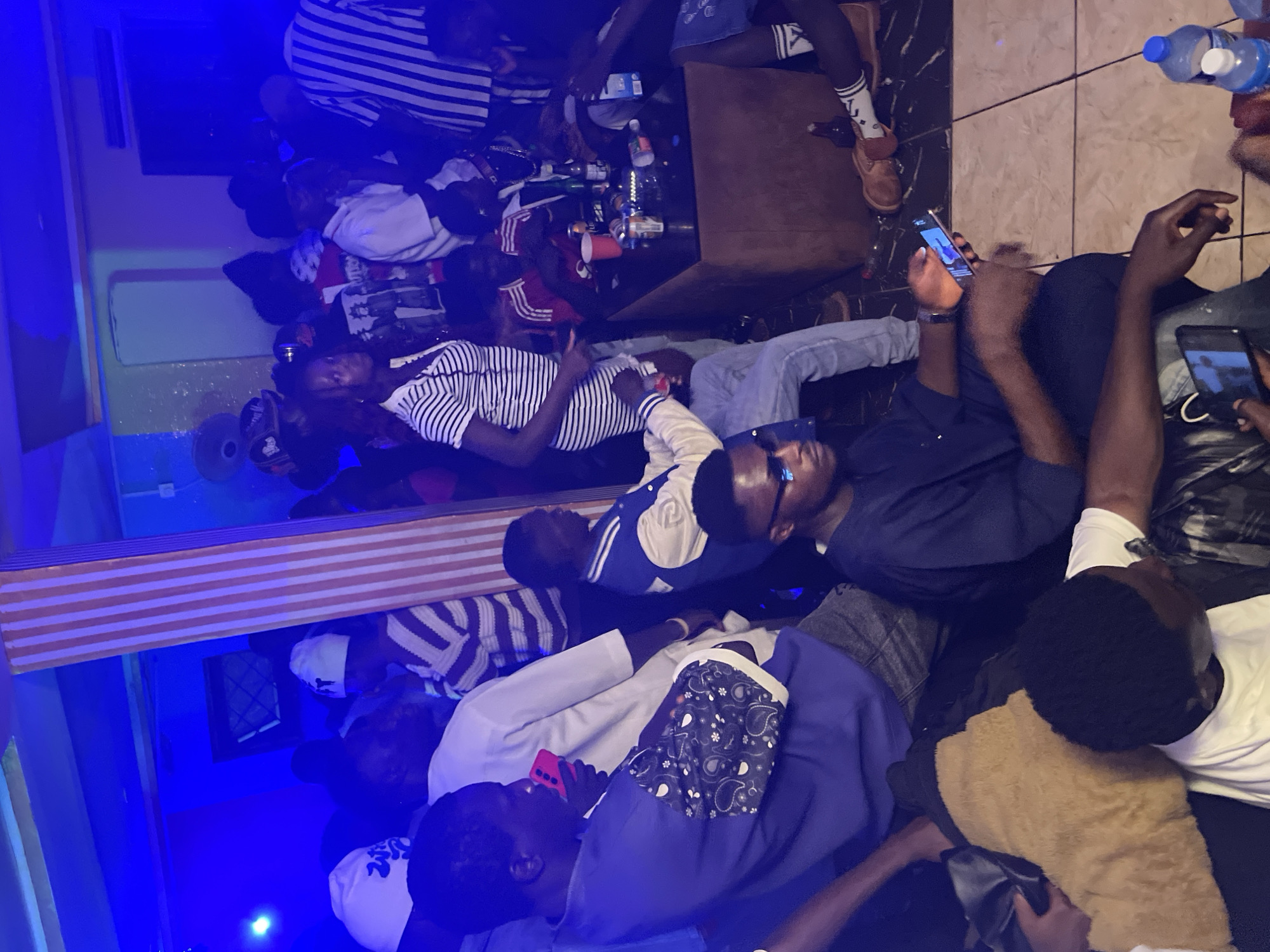 In Wumat, Nigeria, a group of people are gathered indoors in what appears to be a dimly lit lounge or club. Blue lighting illuminates the scene, with a prominent striped pillar in the foreground dividing the space. Several individuals are seated, some looking at their phones, while others are engaged in conversation. A table in the mid-ground holds bottles, cans, and red cups, suggesting a social gathering involving drinks. The attire of the people varies, with casual clothing and headwear being common. The overall atmosphere suggests a relaxed and informal social event.