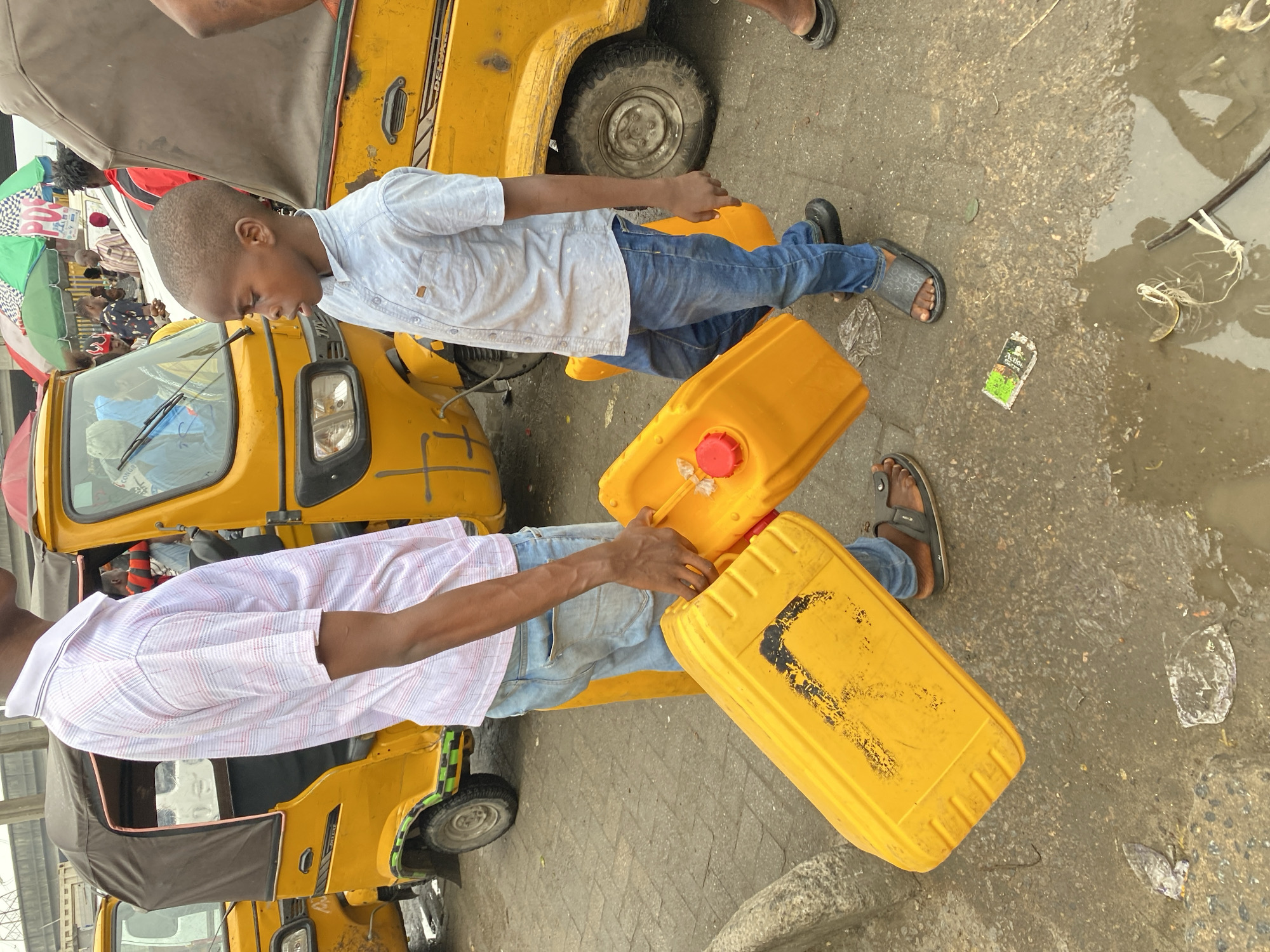Two people are carrying yellow jerrycans on a street in Lagos, Nigeria. The first person is a child, and the second is an adult. Both are wearing sandals and jeans. They are standing next to yellow auto rickshaws. The road is paved, and there is pooled water near the edge of the road.