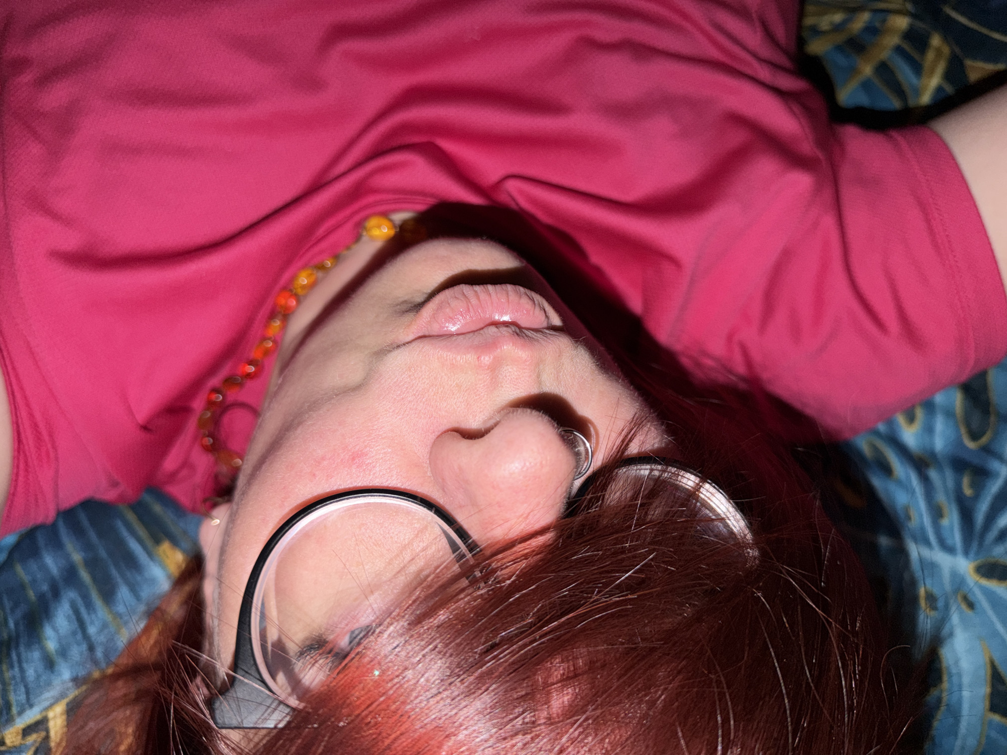 A close-up, angled view of a person with red hair, wearing glasses and a pink shirt. They are lying on a patterned blue and gold surface. A beaded necklace is partially visible. The image provides no information to indicate the location is Chaikovsky, Russia.
