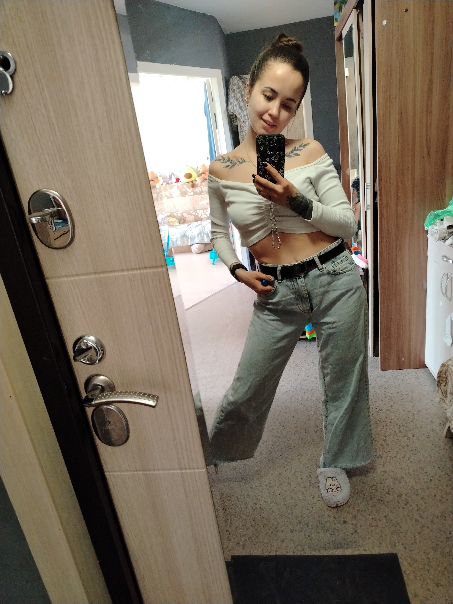 A young woman is taking a mirror selfie in an interior setting. She has a bun hairstyle and is wearing a white off-the-shoulder crop top, light blue jeans with a black belt, and one slipper with a rabbit design. The woman is holding a black smartphone, and tattoos are visible on her collarbones and arm. The reflection shows a room with a glimpse of a bed with soft toys in the background. To the right of the reflection is a wooden wardrobe. The overall environment appears to be a residential room, possibly in Noviy, Russia, though this cannot be definitively confirmed from the image alone.