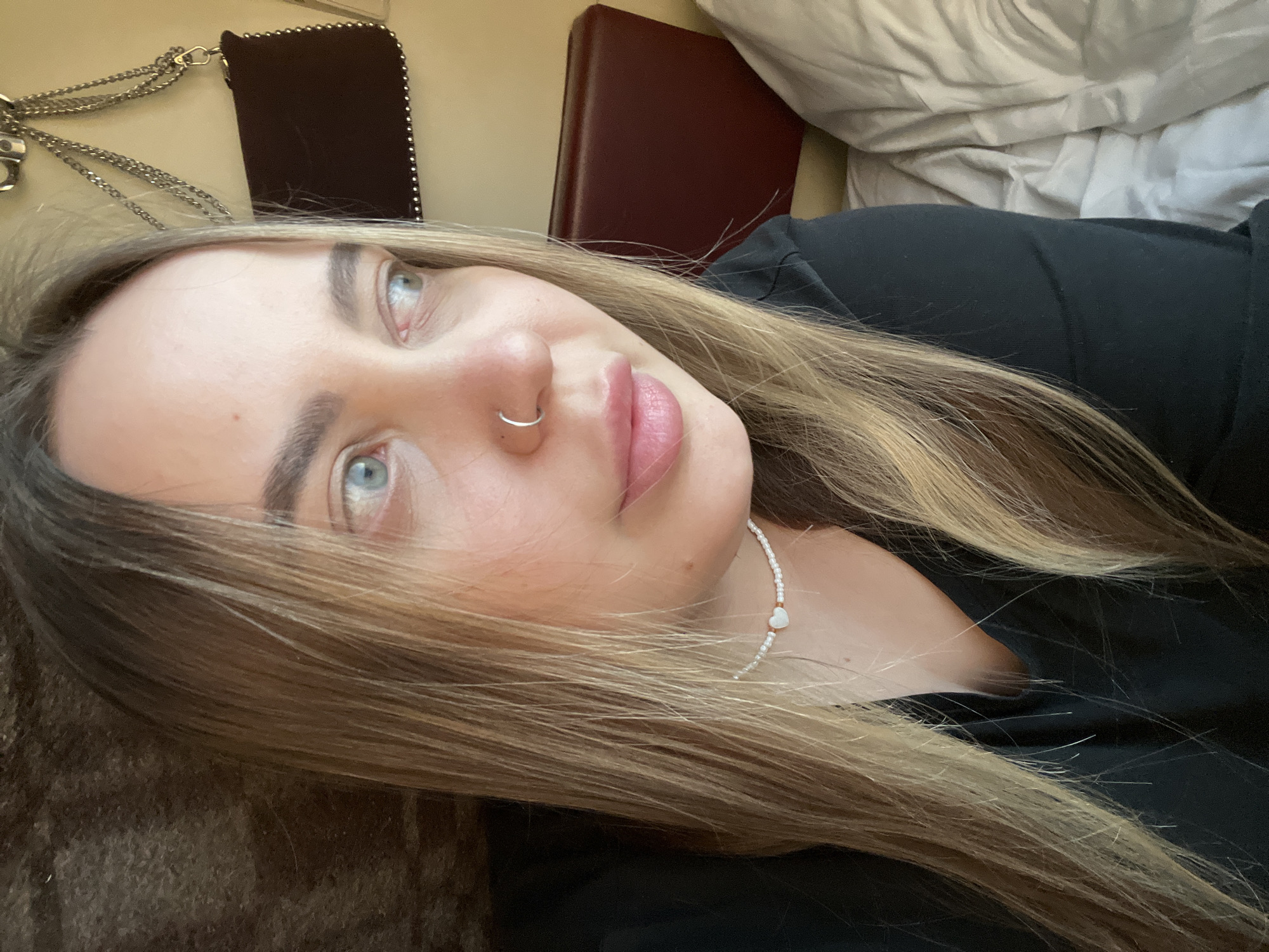 A young woman with fair skin and light brown, layered hair is looking up and to the left. She has blue eyes and a silver nose ring. She is wearing a black shirt and a delicate white beaded necklace with a small heart pendant. The background includes a brown cushioned surface on the left, a dark red cushioned object in the upper center, and a white pillow or duvet in the upper right. A dark-colored purse with a silver chain strap and studded trim is visible in the upper left corner. The photo appears to be taken indoors, possibly in a home. There is no indication of the location being Agryz, Russia.