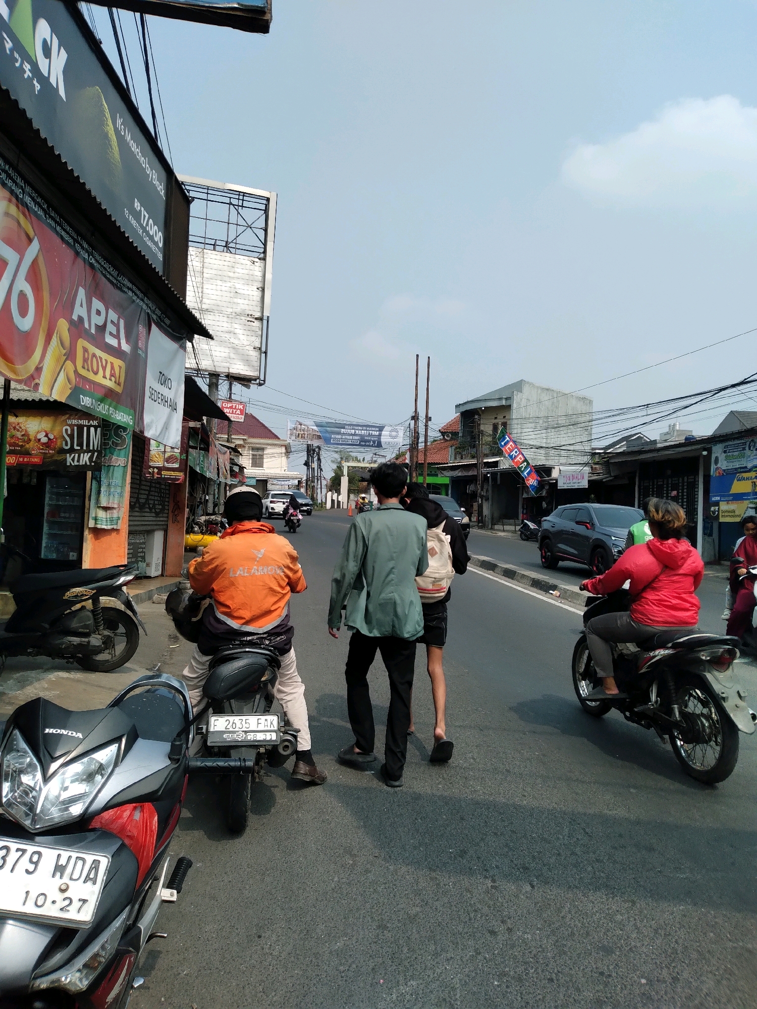 This is a medium-angle, eye-level shot of a street scene in South Tangerang, Indonesia. The foreground is dominated by two motorcycles, one in the lower left and another in the lower center, facing away from the viewer. The motorcycle on the left is primarily black and red with a silver front fairing and a visible license plate that reads "379 WDA 10-27". The motorcycle in the center is black and is being ridden by a person wearing an orange jacket with "LALAMOVE" written on the back. This person is wearing light-colored pants and brown shoes.
In the middle ground, there are several people walking and riding motorcycles. Three individuals are walking away from the viewer towards the right side of the frame. The person on the left is wearing a green jacket and black pants. The person in the middle is wearing a black hoodie and carrying a light-colored backpack. The person on the right is wearing a red jacket and dark pants, and is riding a motorcycle. This motorcycle is black and has a visible license plate that reads "F 2635 FAK".
The background features a street with several buildings lining both sides. On the left, there are shops with various signs, including one that says "76 APEL ROYAL". There are also billboards overhead. The buildings on the right are more residential in appearance, with wires and poles visible above them. The sky is a pale blue with a few scattered clouds. The overall lighting suggests it is daytime, likely late morning or early afternoon, with bright sunlight casting shadows. The scene depicts a typical urban street environment with moderate traffic and pedestrian activity.