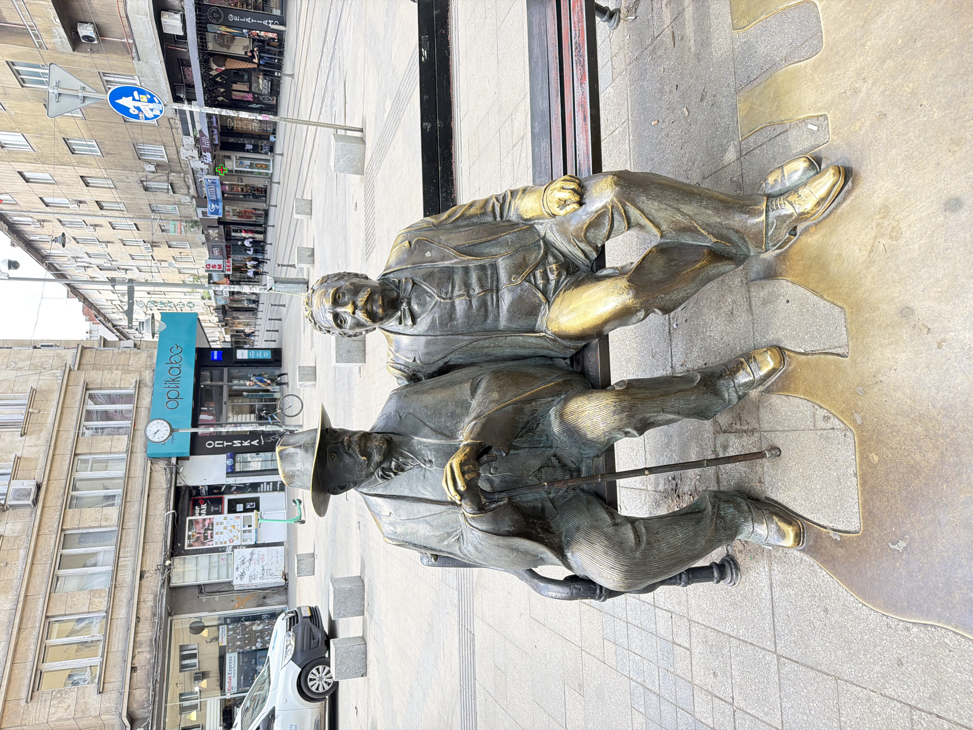 The image shows a bronze sculpture of two men sitting on a bench, located on a pedestrian street in Sofia, Bulgaria. The man on the right has a mustache and is wearing a suit. The man on the left has a hat and a cane. Buildings with shops and apartments are visible in the background. A road sign can be seen on a pole. A white car is parked nearby.