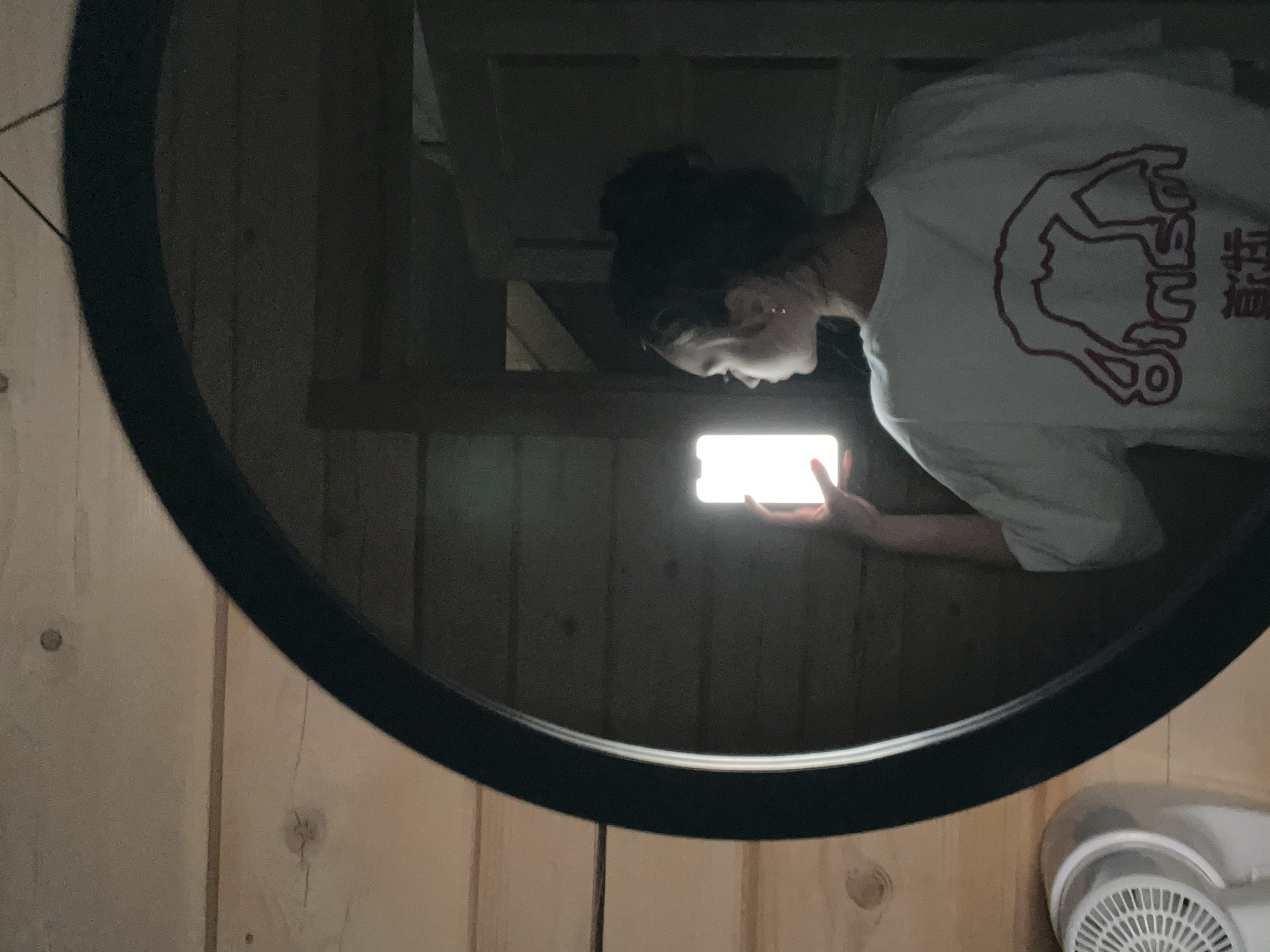 In an indoor setting with wood-paneled walls, a young woman stands in front of a large, round mirror. Her reflection is visible, holding a brightly lit smartphone up to her face. She is wearing a white t-shirt with a red graphic on the back, which appears to be an abstract depiction of a person in a yoga pose with text underneath. Her hair is styled in a bun. To the left, a white appliance, possibly a fan or hair dryer, is partially visible. The lighting in the room is dim, with the smartphone screen serving as a primary light source in the reflection. The overall atmosphere suggests a casual, possibly private moment. The location is not specified in the city of Unknown, Unknown.