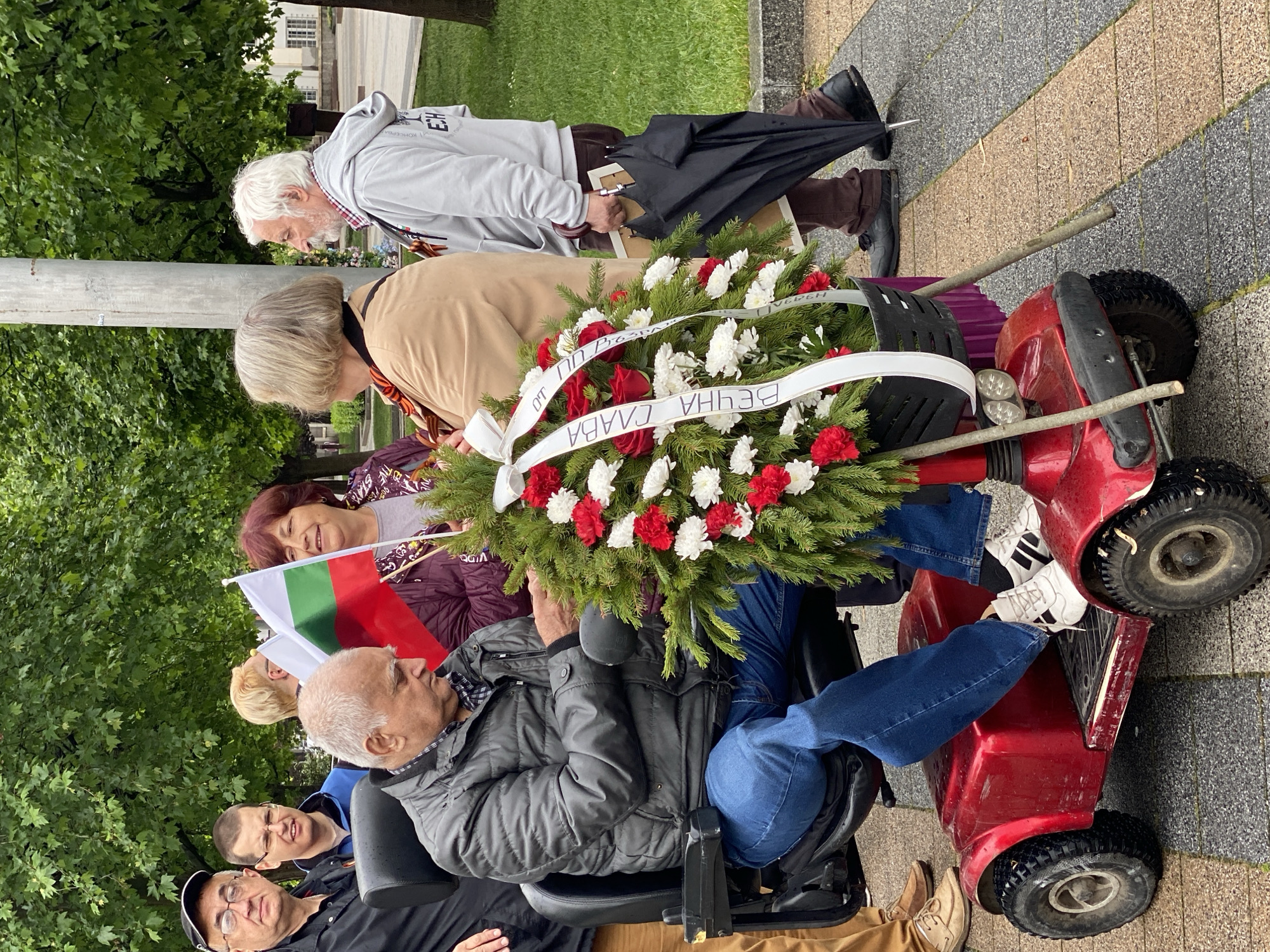 In Pleven, Bulgaria, a group gathers in what appears to be a memorial or commemorative event. An older man is seated in a red mobility scooter, holding a large wreath decorated with red and white flowers and a banner inscribed with Cyrillic text. Several people stand around the scooter and wreath, including a woman holding a Bulgarian flag. Behind them, other individuals are present, some looking down towards the wreath. The setting is outdoors, with trees and a paved area visible in the background, as well as a lamppost.