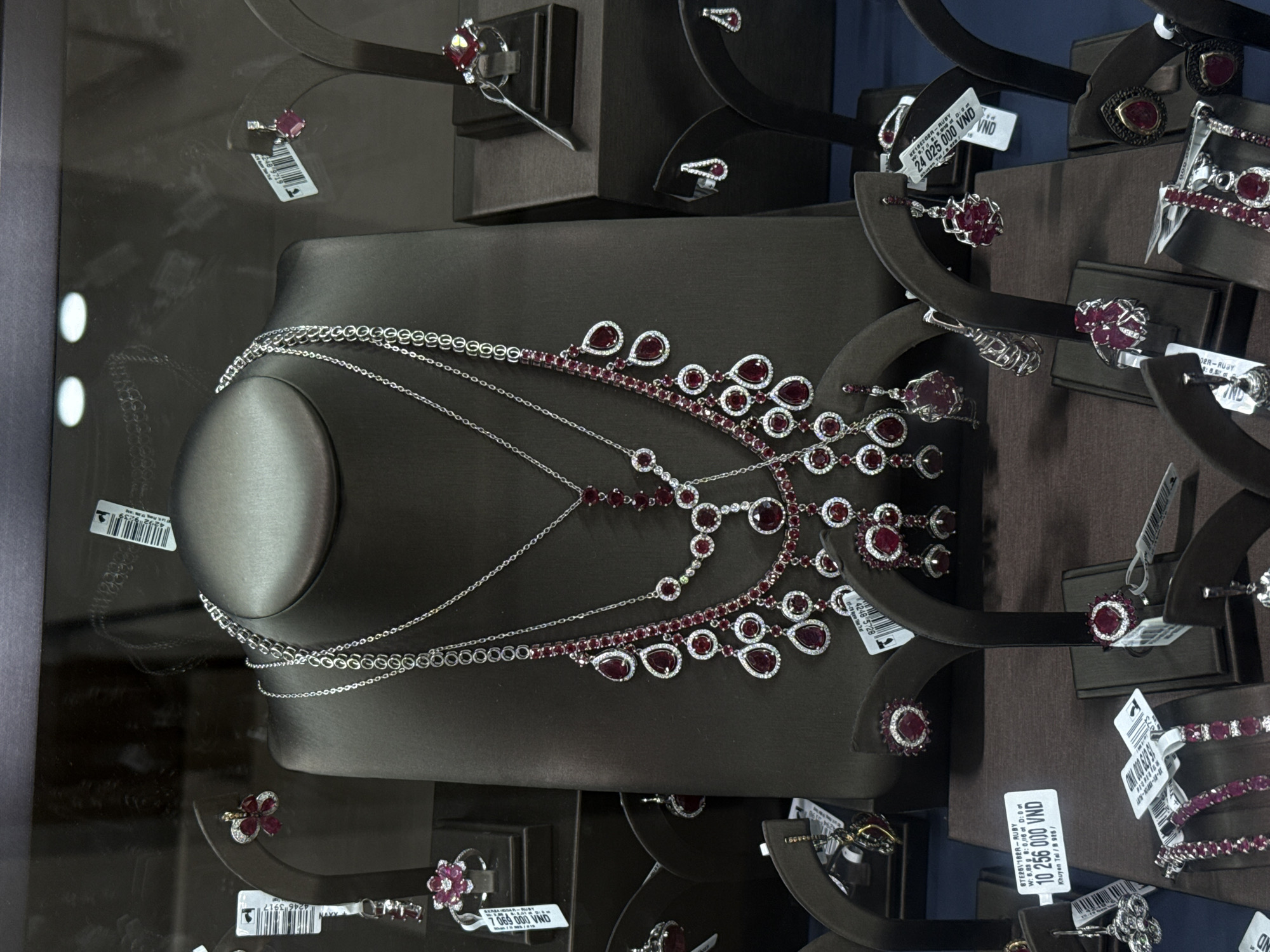 This image shows a display case filled with jewelry, including necklaces, rings, bracelets, and earrings, all set on dark-colored display stands. The jewelry pieces feature a combination of silver metal, diamonds, and prominent red stones, likely rubies. Many of the items have price tags attached, indicating they are for sale. Some tags show a VND currency value. The reflections on the glass of the display case blur the details of the city of Nha Trang Ward, Vietnam location.
