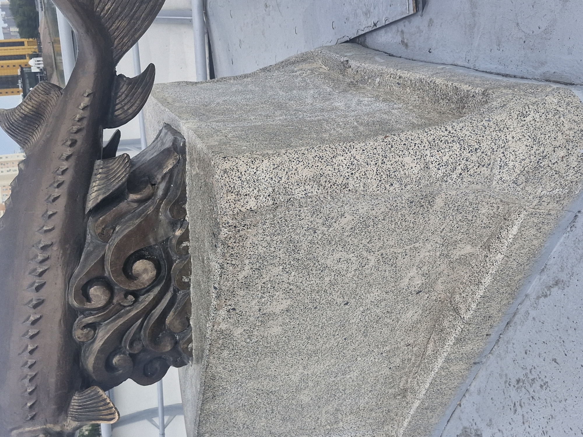 The image shows a bronze sculpture of a fish on a granite pedestal. The sculpture depicts a sturgeon with detailed fins and scales, resting on stylized waves. The pedestal is made of speckled granite and has a multi-faceted design. In the background, a railing and buildings can be seen, suggesting an outdoor urban setting. The image's context indicates it is located in Astana, Kazakhstan. The sculpture is positioned outdoors, likely in a public space such as a park or embankment. No people are visible in the frame. The overall environment appears to be a paved area adjacent to the sculpture.
