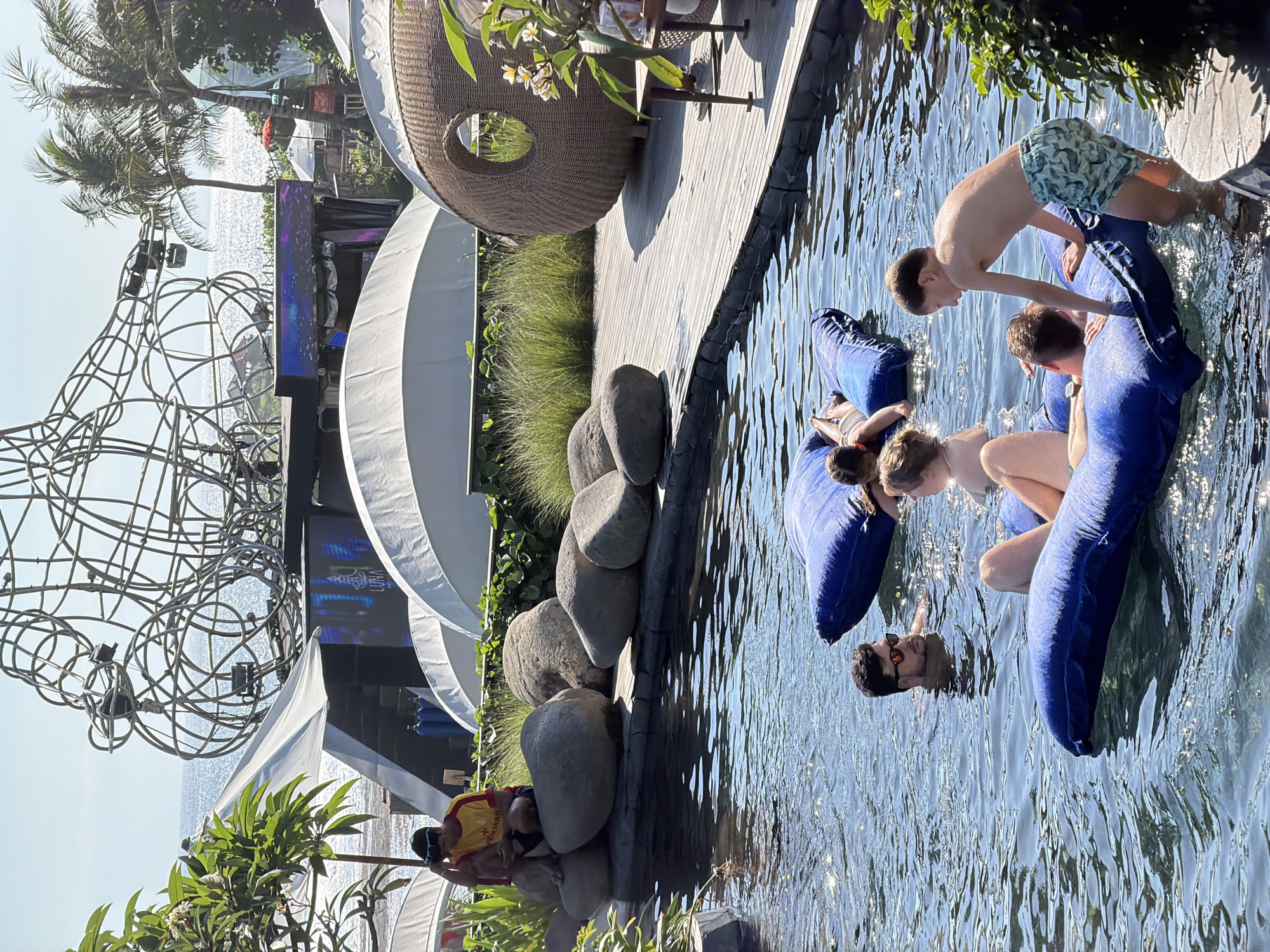This is a photograph taken in Kediri, Indonesia, during the daytime. The scene depicts a group of people enjoying a swimming pool, which appears to be part of a resort or entertainment venue. The pool is large and filled with clear, shimmering water. Several individuals are in the water, some on large inflatable blue rafts, suggesting a leisurely and fun atmosphere.
In the foreground, a young boy in patterned swim shorts is leaning over the edge of a raft, looking into the water. Beside him, two other individuals are partially visible on the same raft. Further to the right, a man with sunglasses is floating in the water, his head just above the surface. Another person is visible in the distance, also in the water.
The background of the image reveals a lush tropical setting. Palm trees and other greenery are visible, indicating a warm climate. There are also elements of resort architecture, including a large woven structure that resembles a lounge chair or pod, and white canopies or tents. To the left, a metal sculpture or structure with a complex, curvilinear design stands prominently, and behind it, a large screen displays what appears to be a performance or event with the word "LUNA" visible. Large, smooth rocks are placed along the edge of the pool, adding a natural aesthetic to the poolside.
The overall mood of the photograph is relaxed and joyful, with people enjoying the water and the sunny weather. The sunlight creates reflections on the water's surface, adding a dynamic element to the scene.