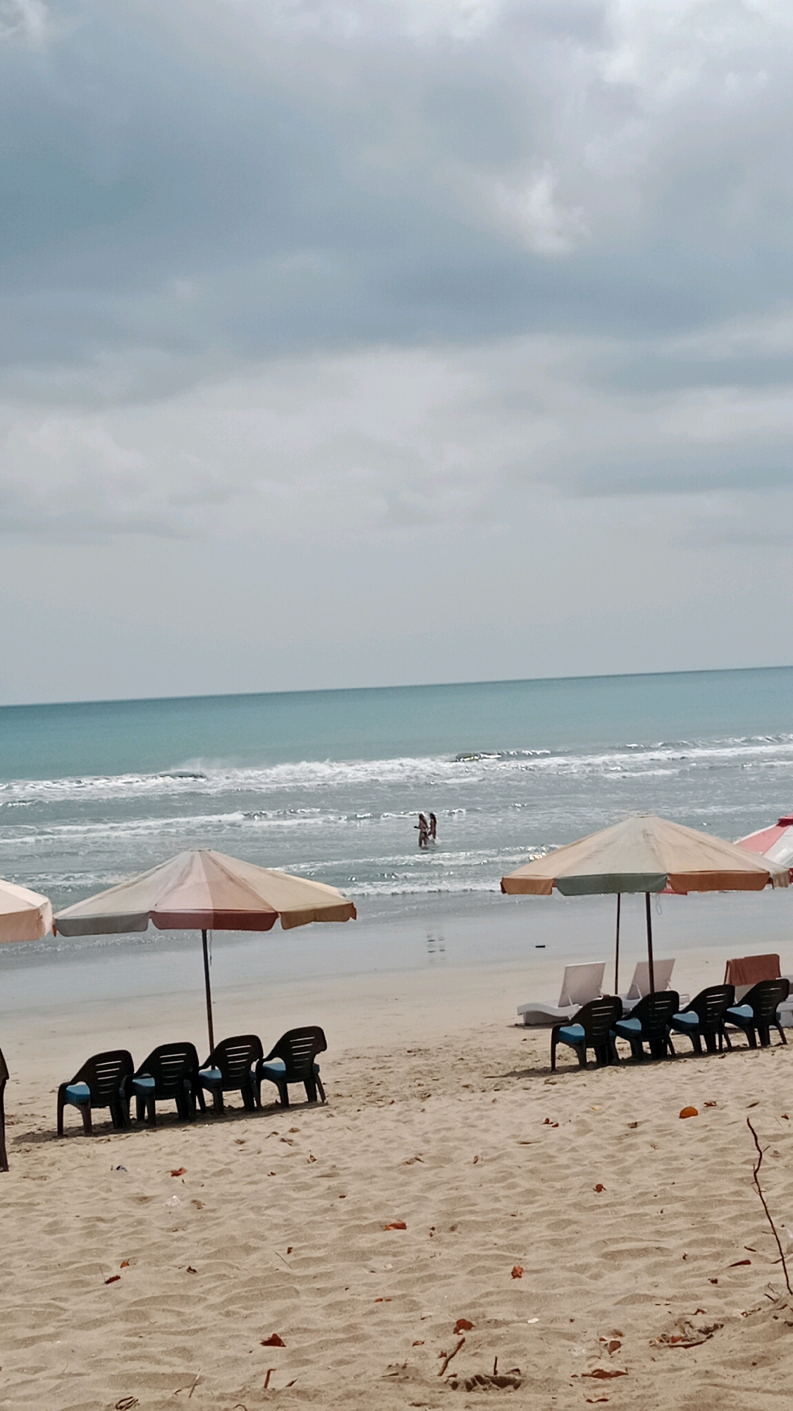 The image depicts a view of Kuta Beach in Indonesia under a cloudy sky. In the foreground, a sandy beach is dotted with scattered leaves and debris. Several empty black lounge chairs with blue cushions are arranged on the sand, some under colorful beach umbrellas. In the middle ground, the ocean features gentle waves breaking near the shore. Two individuals are visible in the water. The background consists of the horizon where the sea meets the overcast sky.