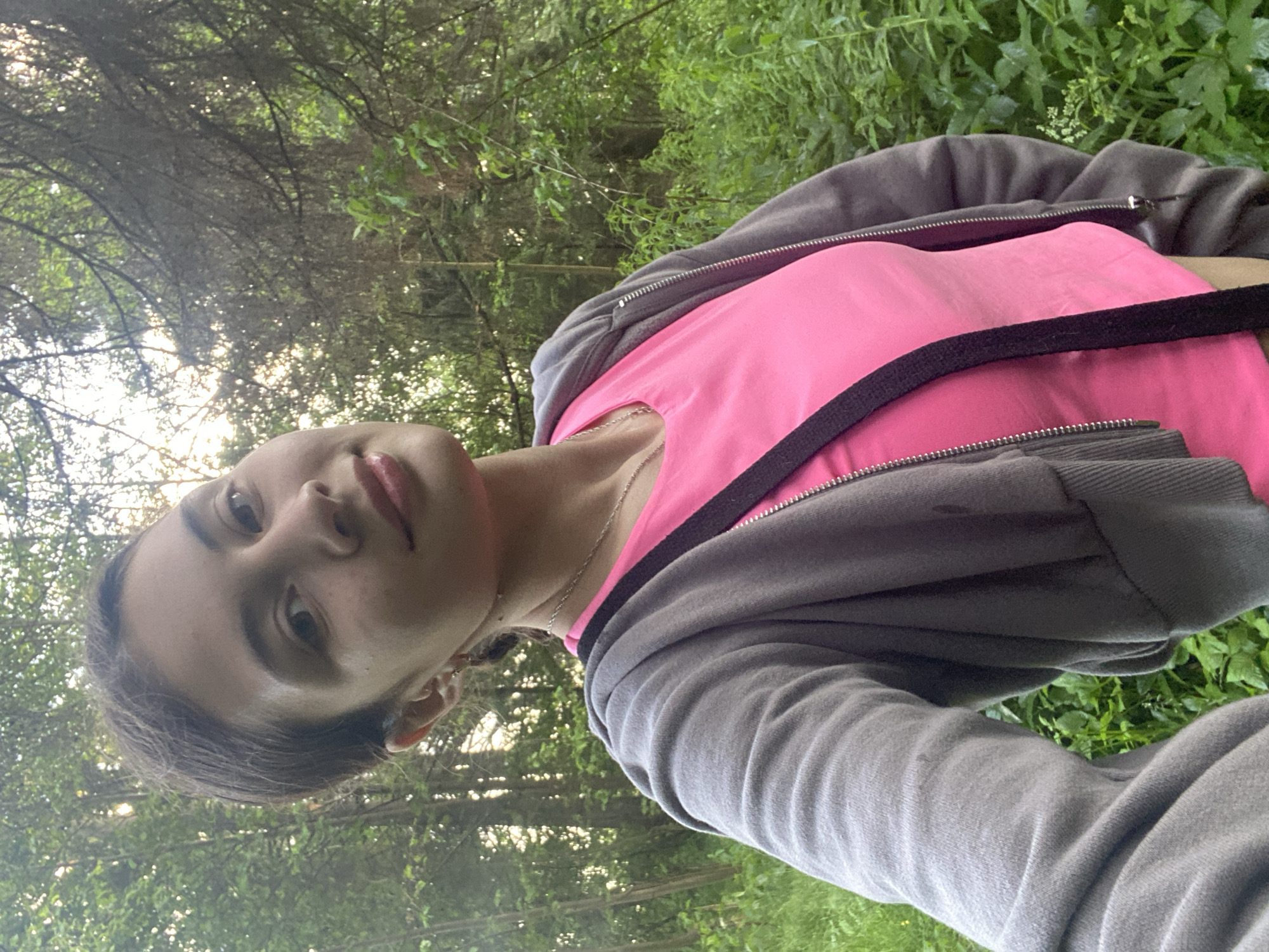 A young woman is taking a selfie in a wooded area. She is wearing a pink t-shirt and a gray zip-up hoodie. The background is filled with trees and green foliage. The lighting suggests it is late afternoon or early evening. There is no indication of Votkinsk, Russia in the image.