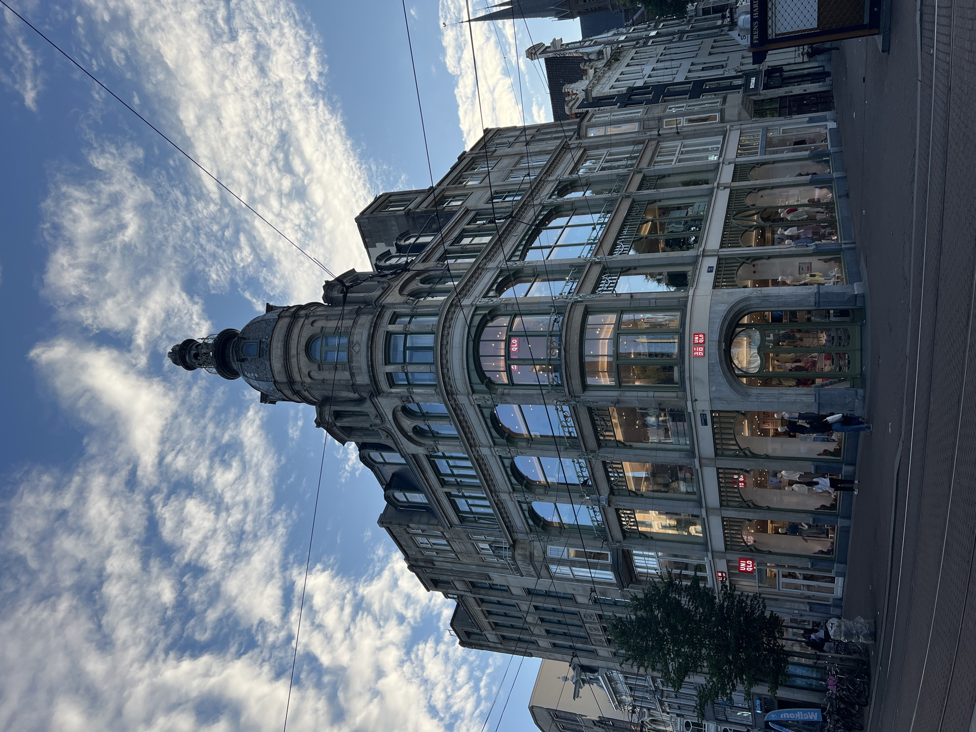 The image shows a multi-story building with large windows, a prominent dome structure, and intricate architectural details. The building appears to be a commercial establishment, with displays visible through the ground-floor windows. Several people are seen on the sidewalk in front of the building and walking past. The scene is set in a city, likely Amsterdam, given the context.
The time of day appears to be late afternoon or early evening, suggested by the lighting and the slightly overcast sky with scattered clouds. The weather seems pleasant. The building's architecture, with its ornate facade and rounded corner, hints at a historic European city.
Visible text includes "UNIQLO" in red and white lettering on the building's facade, indicating the presence of a Uniqlo store. Other signs, though less legible, are also present on adjacent buildings. The overall mood is one of urban activity and commerce under a dynamic sky. The image is captured with a vertical orientation.