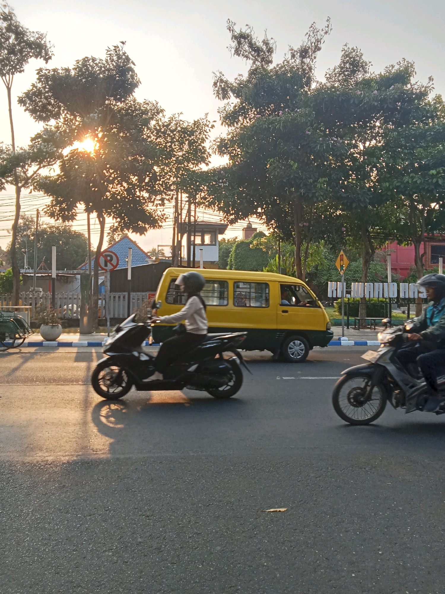The image captures a street scene in Kaliwungu, Indonesia, during what appears to be sunset. The foreground is dominated by an asphalt road, reflecting the warm, low-angled sunlight. Two motorcycles are in motion. On the left, a black motorcycle is ridden by a person wearing a light-colored long-sleeved shirt and dark pants, with a black helmet. The motorcycle's shadow is cast on the wet-looking road. On the right, another motorcycle, possibly silver and black, is ridden by a person wearing a dark jacket and helmet. In the mid-ground, a yellow and green van is parked or moving slowly. Behind the van, trees line the street, with the sun partially visible through their branches, creating a lens flare effect. A "No Parking" sign with a red circle and a diagonal bar over a "P" is visible on the left side of the road. Further in the background, there are buildings, some with distinctive blue roofs, and more trees. A traffic signal is mounted on a pole in the distance. The overall atmosphere suggests a typical urban environment during golden hour.