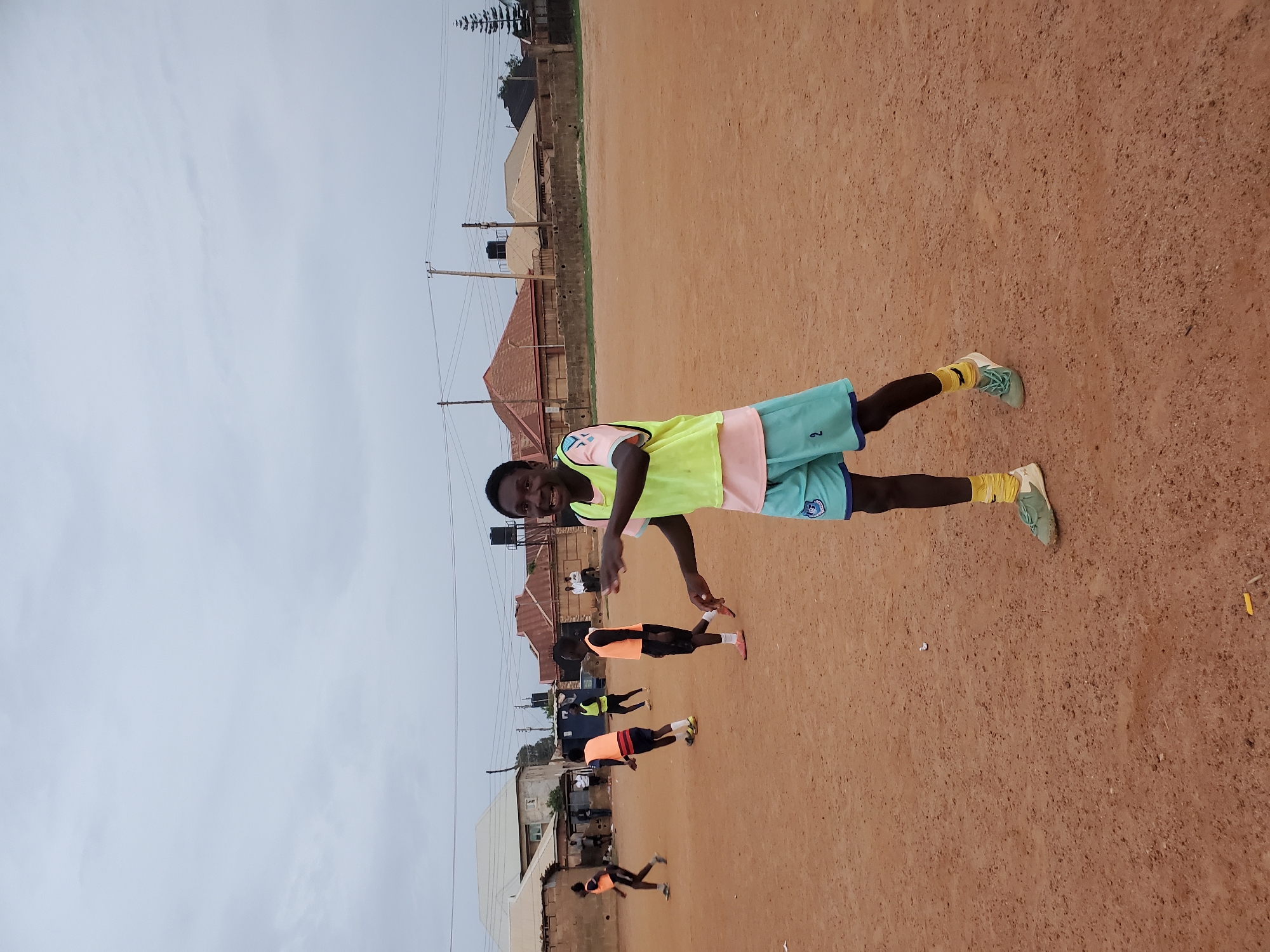 The image depicts a young man in a sports jersey and shorts standing on a dirt field. He is smiling and looking directly at the camera. In the background, several other people are engaged in athletic activity on the same field. Residential buildings and utility poles are visible in the distance. The sky is overcast. Based on the architecture and general environment, the location is likely in Jos, Nigeria.