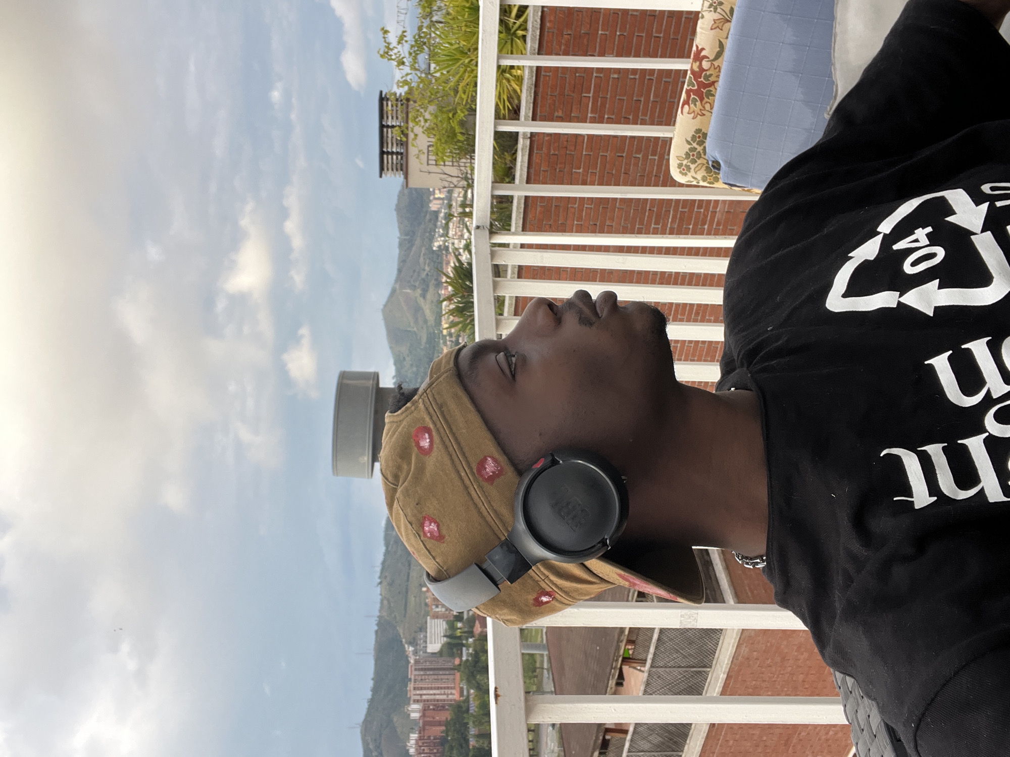A medium shot depicts a Black man looking upward toward the sky. He wears a black t-shirt with white lettering, a tan cap with red designs, and black headphones. The location appears to be a balcony or rooftop overlooking a city, possibly Barcelona, Spain, judging by the urban landscape with mountains in the background. The sky is partly cloudy.