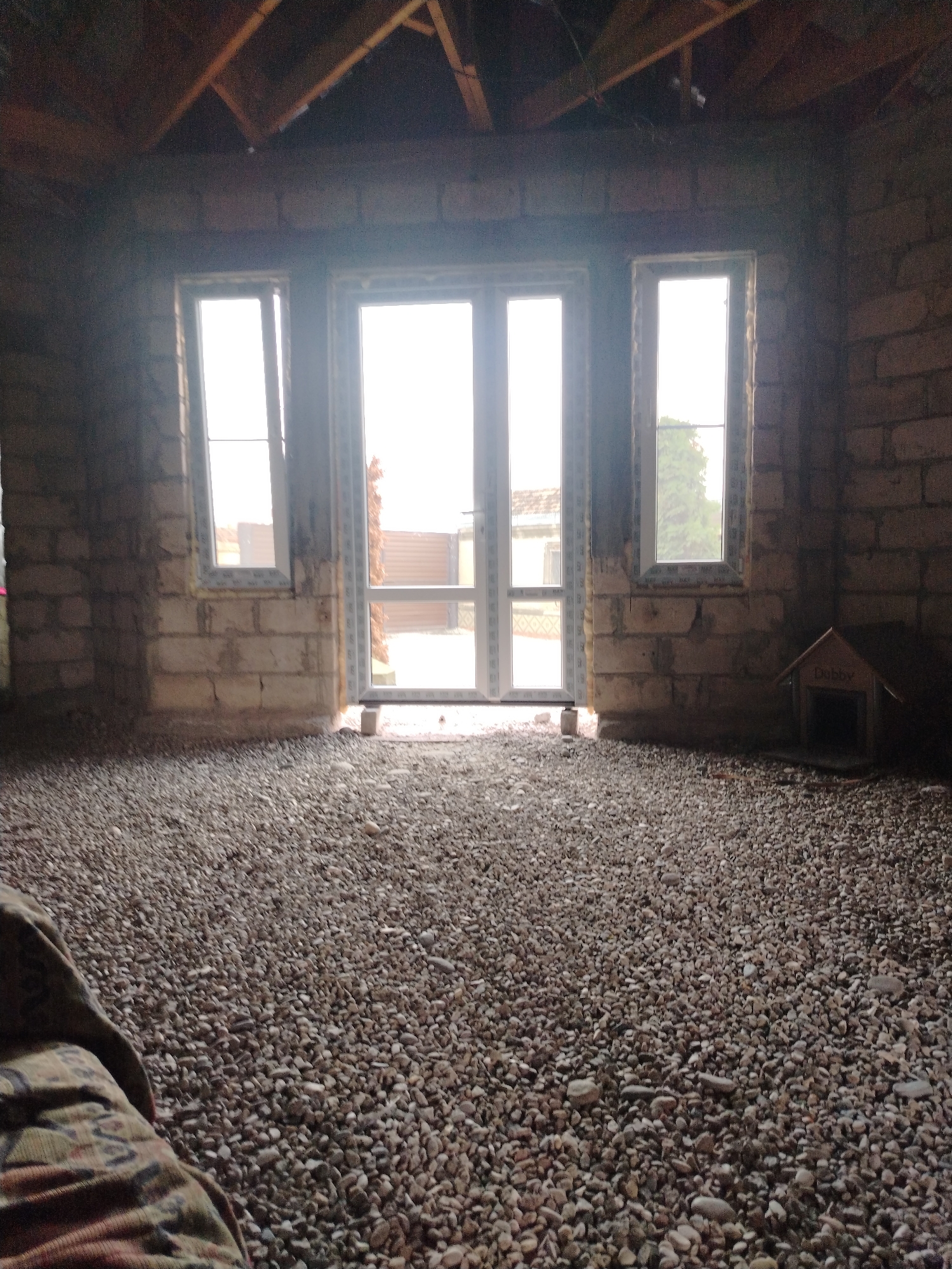 The image depicts the interior of an unfinished building with exposed wooden rafters visible on the ceiling. The walls are constructed of light-colored bricks. The floor is covered with a thick layer of small, light-colored pebbles. In the center of the room is a large, double glass door, flanked by two tall, narrow windows. Through the glass doors, an outdoor scene is visible, including a corrugated metal fence and some trees. To the right of the doors, a small, wooden dog house with "Dobby" written on it is placed on the pebble floor. A portion of a patterned fabric, possibly a blanket or upholstery, is visible in the lower left corner of the image. The lighting suggests it is daytime, with bright light coming from the doors and windows. The location is stated to be in the city of Атажукино, Russia.