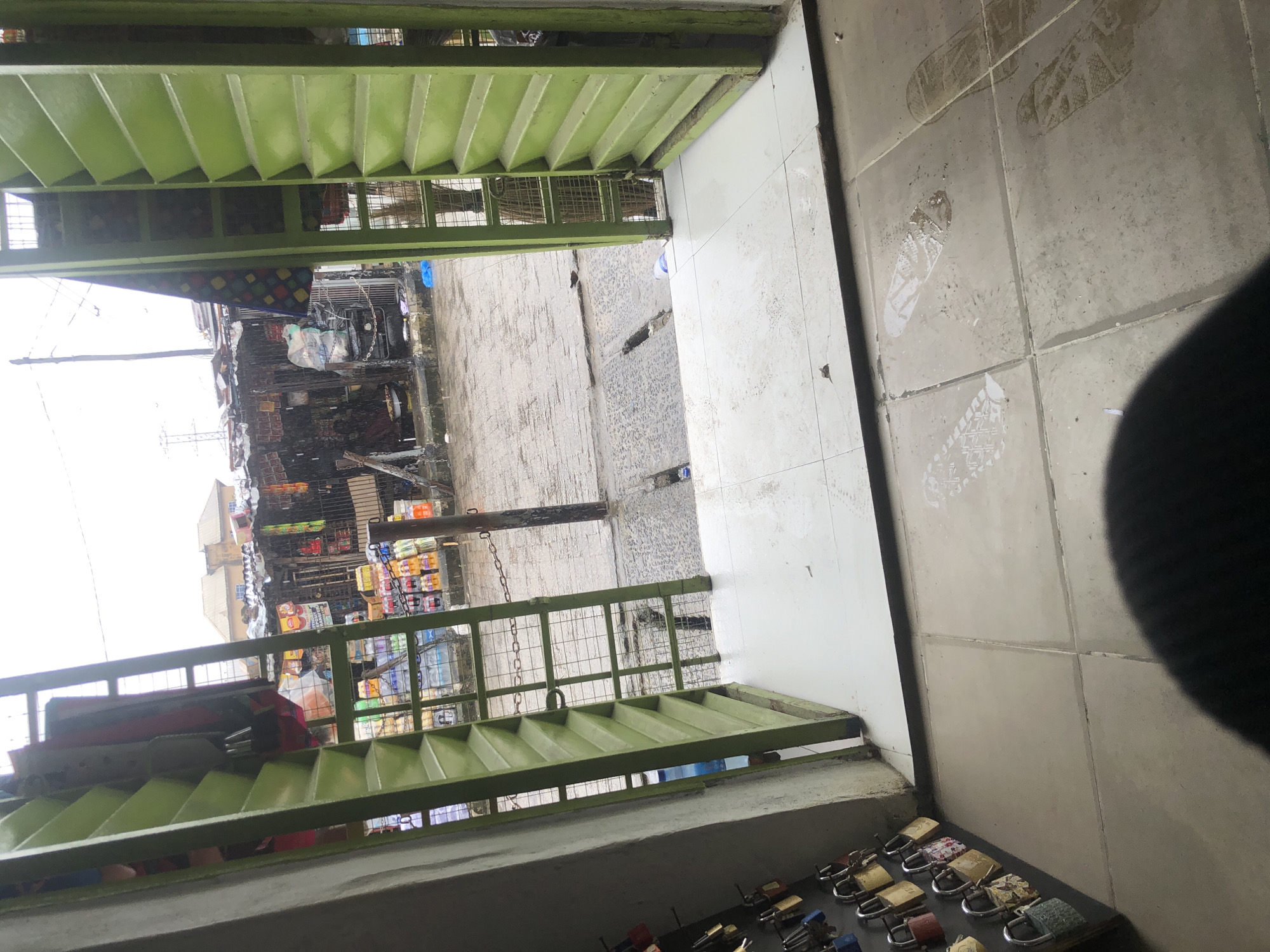 The image captures an outdoor scene in Ado, Nigeria, looking through a green metal grate with horizontal louvers. The foreground consists of a tiled floor with several visible shoe prints, some appearing wet. To the left, a wall is lined with numerous padlocks of varying colors and designs, secured to a black surface. Beyond the grate, a street is visible, appearing wet and possibly under light rain. Along the street, a row of makeshift stalls or shops is set up, displaying various goods, including bottled drinks and other merchandise. A chain extends across the street in front of the stalls. In the background, buildings and power lines are faintly discernible. The overall impression is of a bustling, possibly market-like environment in a Nigerian city, under overcast or rainy weather conditions.