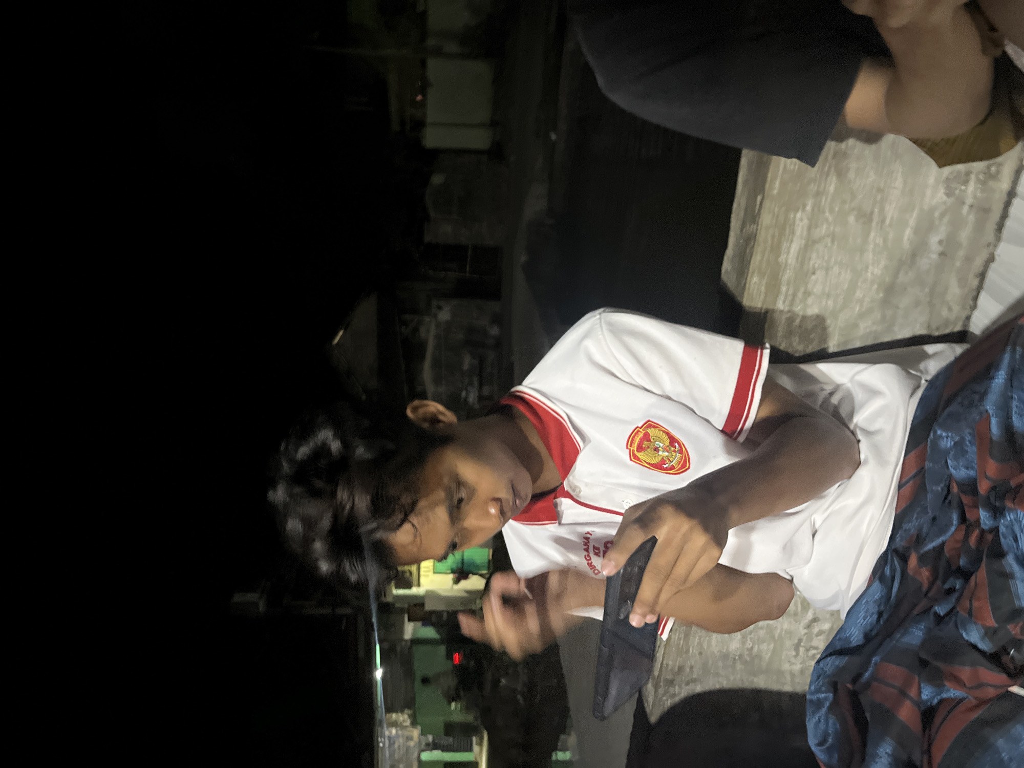 An Indonesian male teenager, wearing a white polo shirt with red trim and an emblem, is seated at night, engrossed in using a smartphone. He is positioned on a textured, light-colored surface that appears to be a concrete bench or ledge. To his left, a dark blue and red striped fabric is visible, draped over his lap. In the background, out-of-focus structures and greenery suggest an outdoor setting within a residential area. The general ambiance is that of a dimly lit street or alleyway in Laban, Indonesia. Another individual, partially visible on the right, is also seated, wearing a black t-shirt. The image captures a moment of leisure, possibly amidst a community gathering or a casual hangout in the evening.
