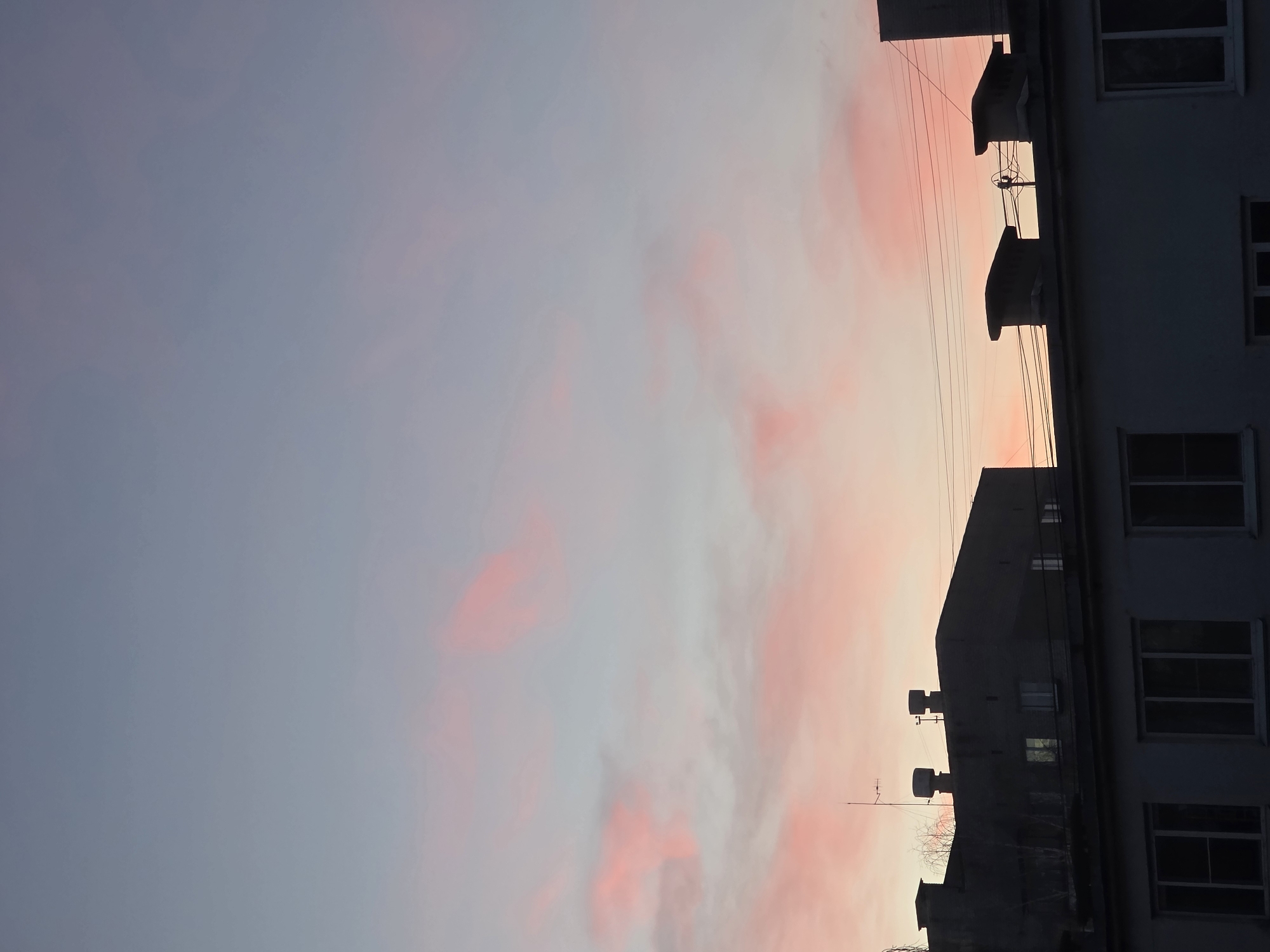 The image is a photograph taken in the city of Chaikovsky, Russia. It shows a sunset sky with pink and white clouds, juxtaposed against the silhouette of a multi-story building on the right side of the frame. The building appears to be residential, with visible windows and architectural features such as balconies and chimneys. Power lines run across the sky in front of the building.