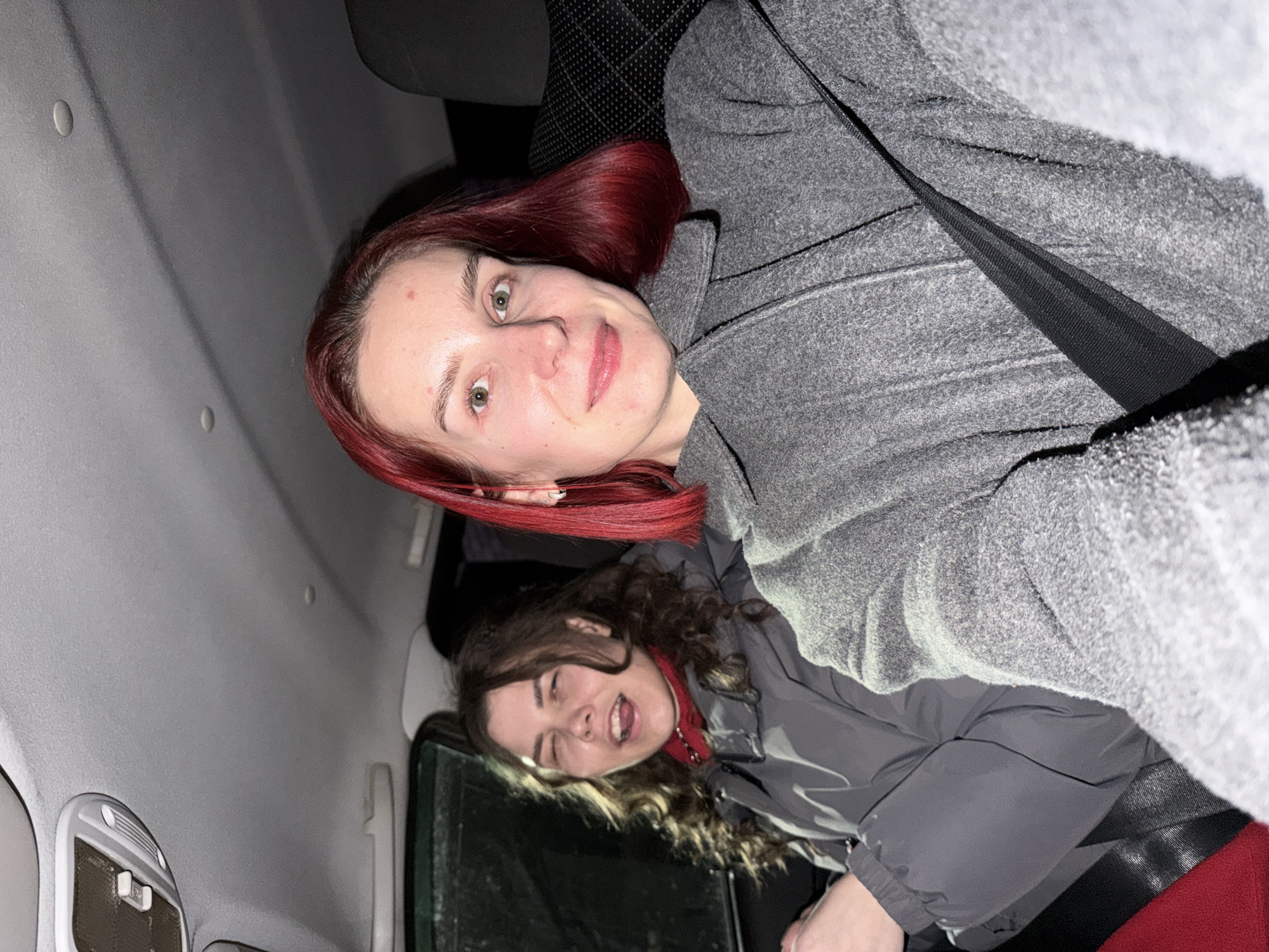 The media file captures two young women inside the passenger compartment of a vehicle, likely a car, in Chaikovsky, Russia. The scene appears to be set at night or in low-light conditions, indicated by the artificial interior lighting and the dark exterior visible through the window.
In the immediate foreground, a woman with long, reddish-brown hair and light skin smiles gently at the camera, her green eyes wide open. She is wearing a grey, textured coat, and a black seatbelt strap is visible across her shoulder. A small red blemish is noticeable on her forehead.
Behind and slightly to her right, another woman with dark, curly hair is visible. She is dressed in a grey puffer jacket over a red layer, possibly a turtleneck. This woman is making a playful or silly face, with her eyes squinted and mouth open, her tongue slightly visible.
The vehicle's interior features a light grey headliner and dark, patterned seats. A grey interior light fixture is visible on the ceiling of the car. Both individuals appear to be passengers, and their warm clothing suggests a cool climate. No visible text is present in the image.