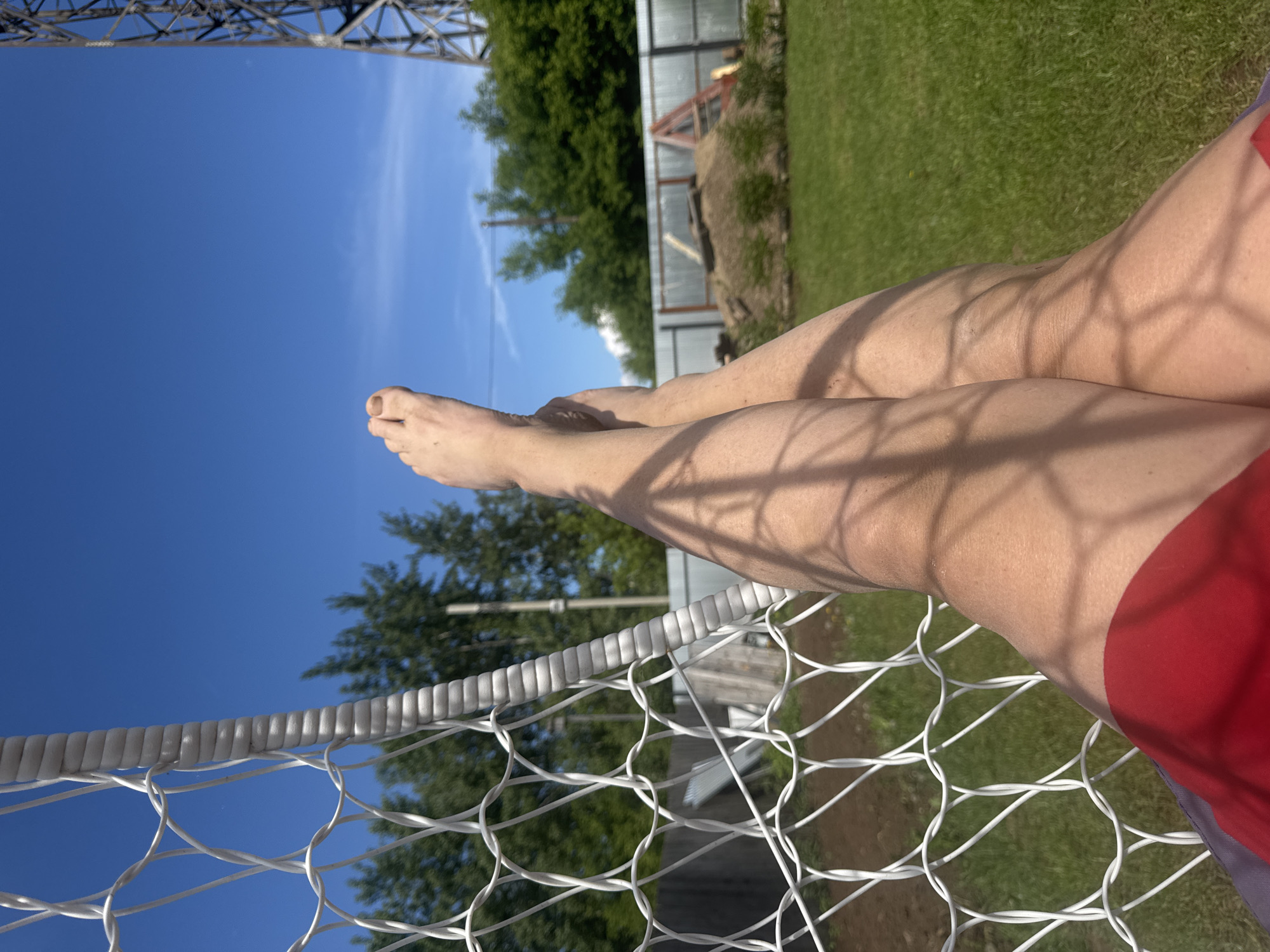 The image captures a first-person perspective of a person's bare legs and feet resting on a white net, likely a hammock or recliner, outdoors in Chaikovsky, Russia. The person is wearing red shorts or swim trunks. Sunlight casts distinct, geometric shadows from the net across their skin, highlighting the sunny conditions.
The scene is set under a clear, vibrant blue sky. In the background, lush green grass is visible, along with a metal fence, dense green trees, and what appears to be a small greenhouse or shed. Part of a large metal structure, possibly a power line tower or a bridge, is also visible against the sky on the left side of the frame. The bright illumination and lack of clouds indicate a sunny day. The activity depicted suggests a moment of relaxation or sunbathing. No visible text is present in the image.