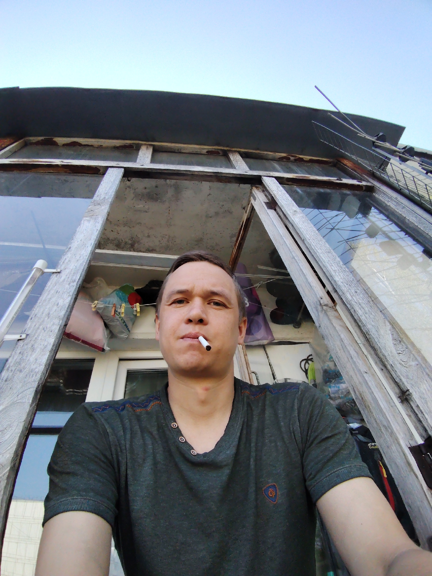 A low-angle, medium shot depicts a man with a cigarette in his mouth, standing in front of an old, wooden-framed window. The window's glass panes are partially visible and reflect the outdoor environment. Behind the man, inside the building or structure, are miscellaneous items. Above the window frame is a dark, flat roof or overhang, and further above that is a pale blue sky. An antenna is visible on the right side of the frame. The man is wearing a dark gray-green t-shirt with a small emblem on the lower right and decorative stitching on the shoulders. The location is Chaikovsky, Russia.