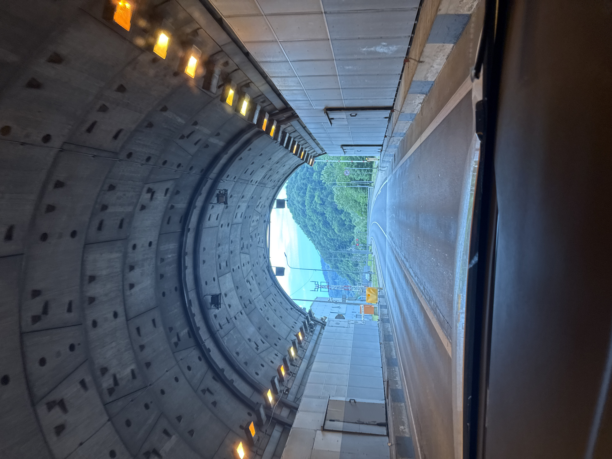 The image shows a perspective from inside a vehicle exiting a tunnel. The tunnel ceiling is constructed from concrete segments, with regularly spaced lights illuminating the interior. The walls of the tunnel are paneled and appear to be made of metal or a composite material. Orange lights are visible on the right side of the tunnel, suggesting illumination. The road surface is wet, indicating recent rain or a damp environment.
As the vehicle approaches the tunnel exit, the scene transitions to an exterior view. Lush green mountains covered in trees dominate the background, with a bright, cloudy sky overhead. A paved road curves to the right, with a white line marking the lane division. Various structures are visible near the tunnel exit, including what appears to be a traffic signal pole with red and white indicators, a yellow barrier, and some industrial-looking buildings or access points along the tunnel wall. A sign with a red circle and a diagonal line, likely indicating a prohibition, is visible on the left side of the road.
The image does not contain any people or explicit actions. The focus is on the infrastructure of the tunnel and the natural landscape at its exit. The location identified is the Kepsha Tunnel in Kepsha, Russia.